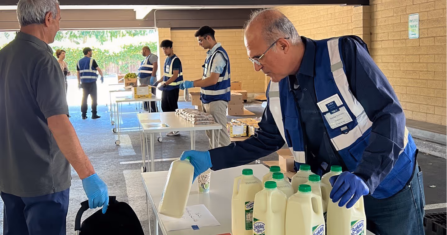 Volunteers wearing blue vests and gloves distributing milk and groceries at an outdoor food assistance site.
