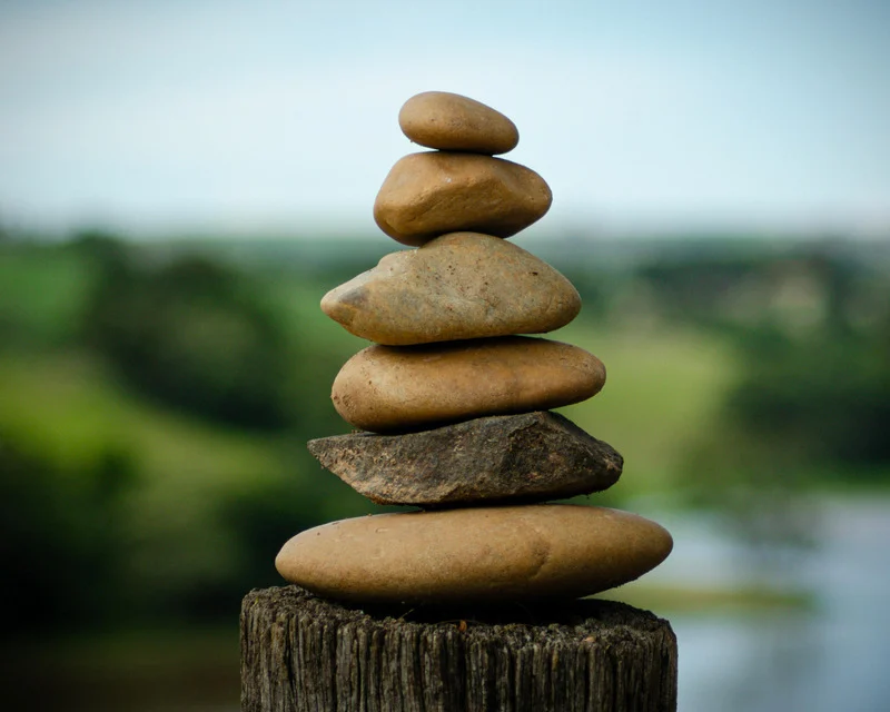 Une pile de six pierres lisses de différentes tailles équilibrées sur un poteau en bois avec un arrière-plan flou de verdure et de ciel clair.