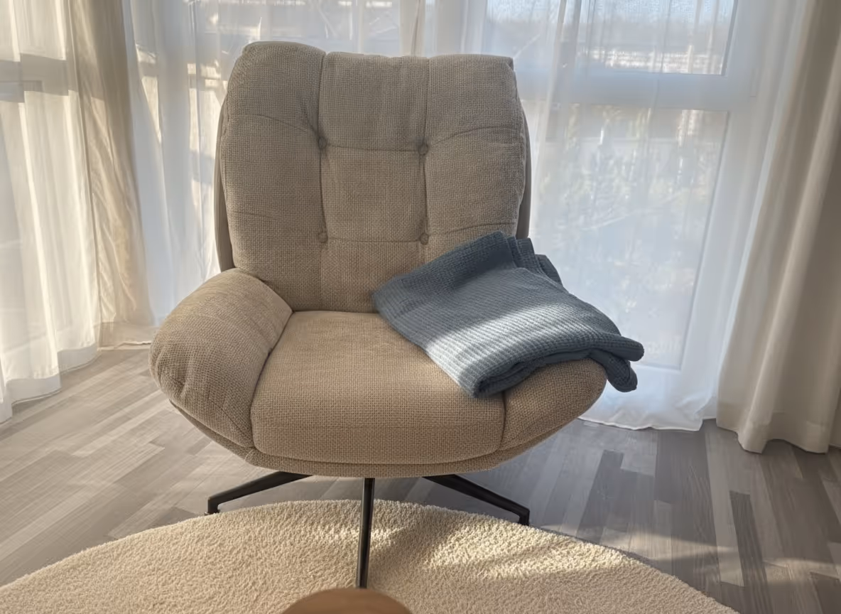 Beiger gepolsterter Sessel mit einem gefalteten grauen Strickpullover darauf vor einem Fenster mit weißen Gardinen.