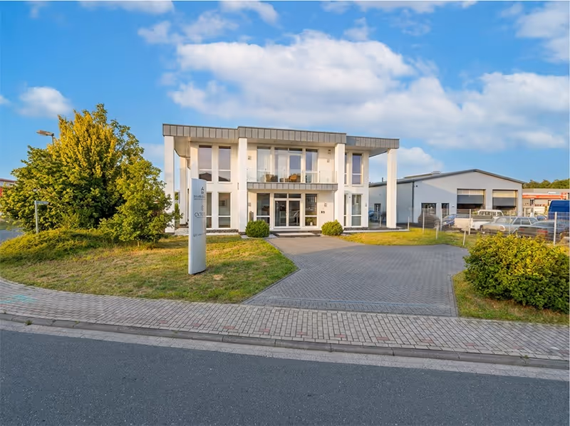 Modernes Bürogebäude mit großem Glasfront-Eingang, gepflasterter Einfahrt und grüner Umgebung unter blauem Himmel.