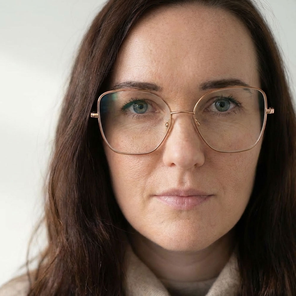 Nahaufnahme einer Frau mit langen braunen Haaren und großen, rechteckigen Brillengläsern mit goldfarbenem Rahmen.