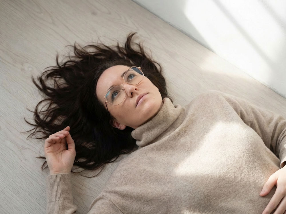 Frau mit Brille und braunem Pullover liegt auf hellem Holzboden und schaut nach oben.
