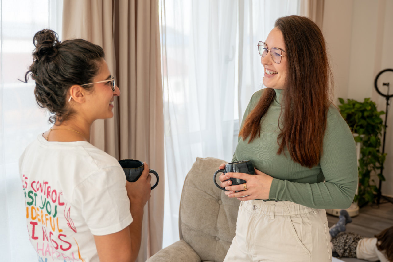 Zwei Frauen mit Brillen unterhalten sich lachend und halten schwarze Tassen in einem hellen Wohnzimmer.
