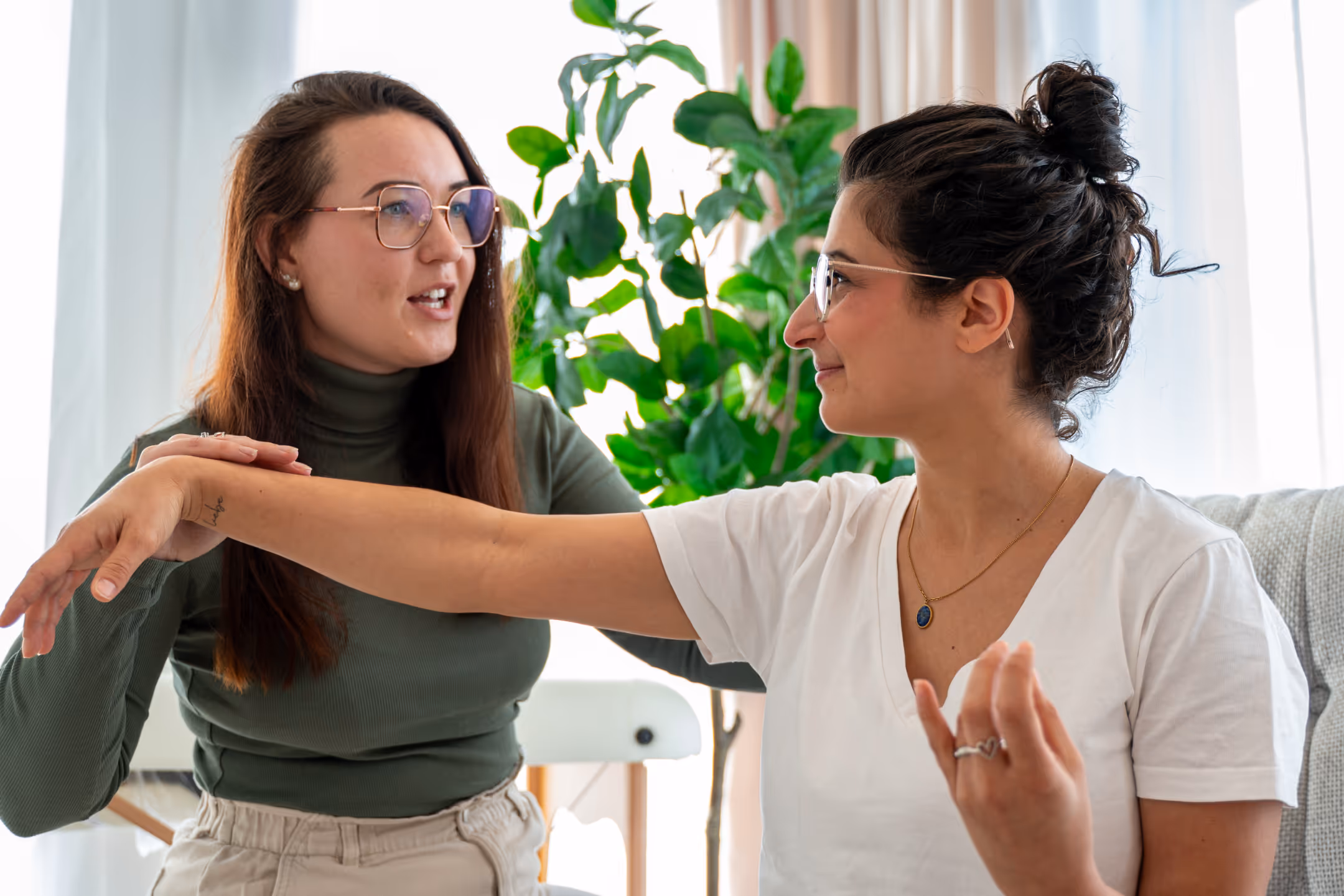 Zwei Frauen unterhalten sich, eine streckt den Arm aus, während die andere ihn hält, im hellen Raum mit grünen Pflanzen im Hintergrund.
