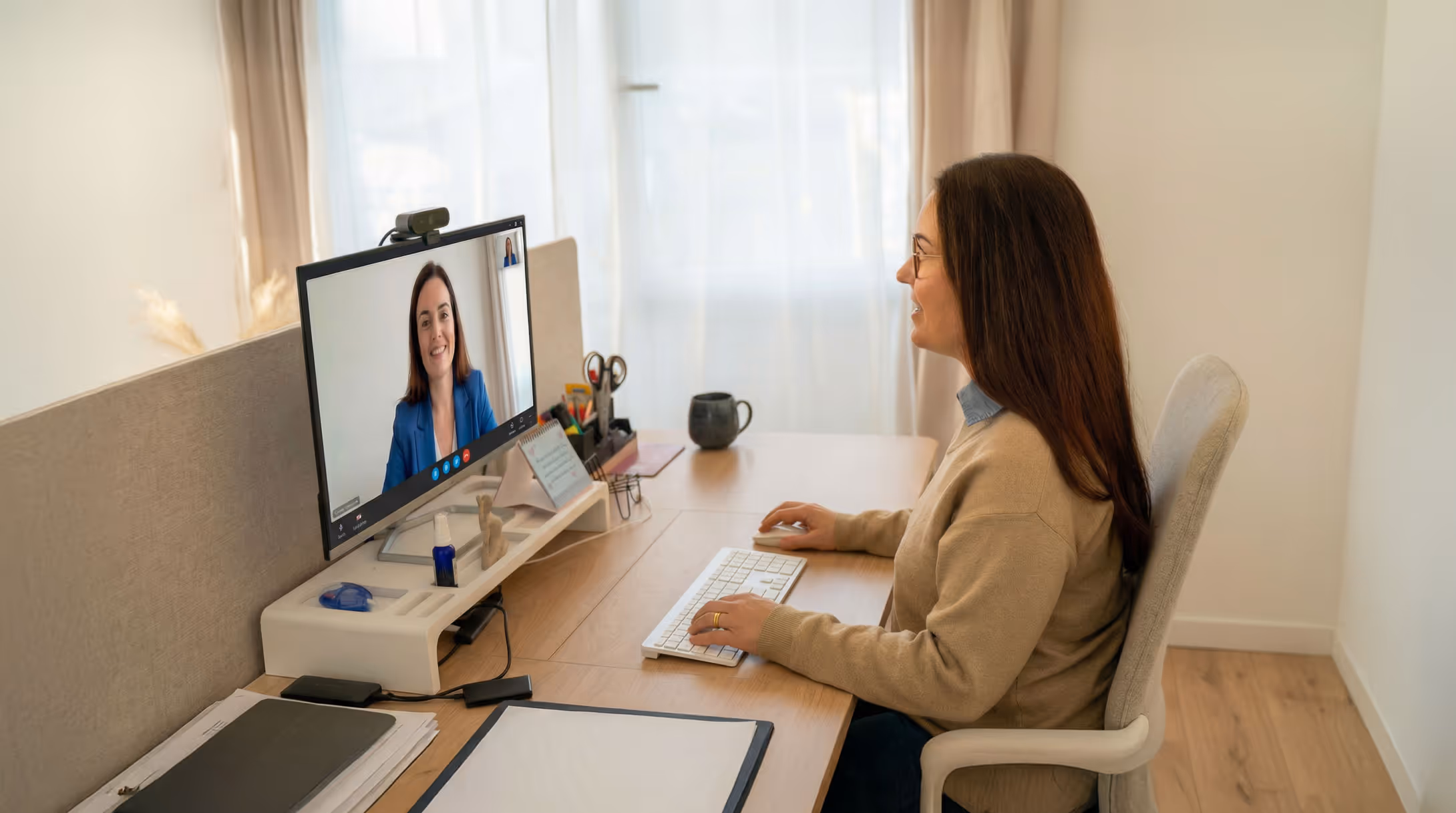Frau mit Brille sitzt an einem Schreibtisch und führt ein Videogespräch mit einer lächelnden Frau auf dem Computerbildschirm.