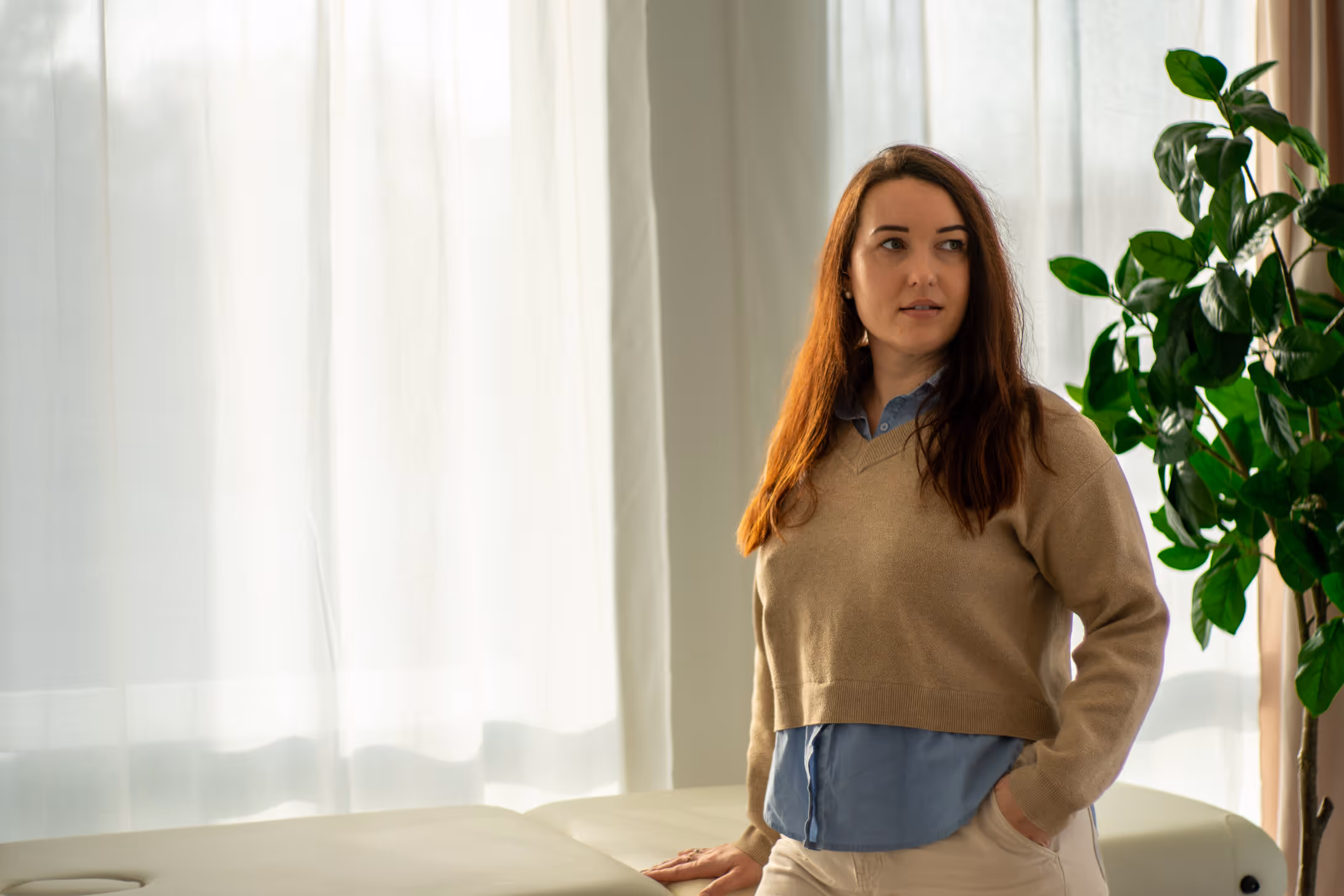 Frau mit braunen Haaren in beigem Pullover lehnt an einem Möbelstück neben einer grünen Topfpflanze vor einem weiß durchscheinenden Vorhang.