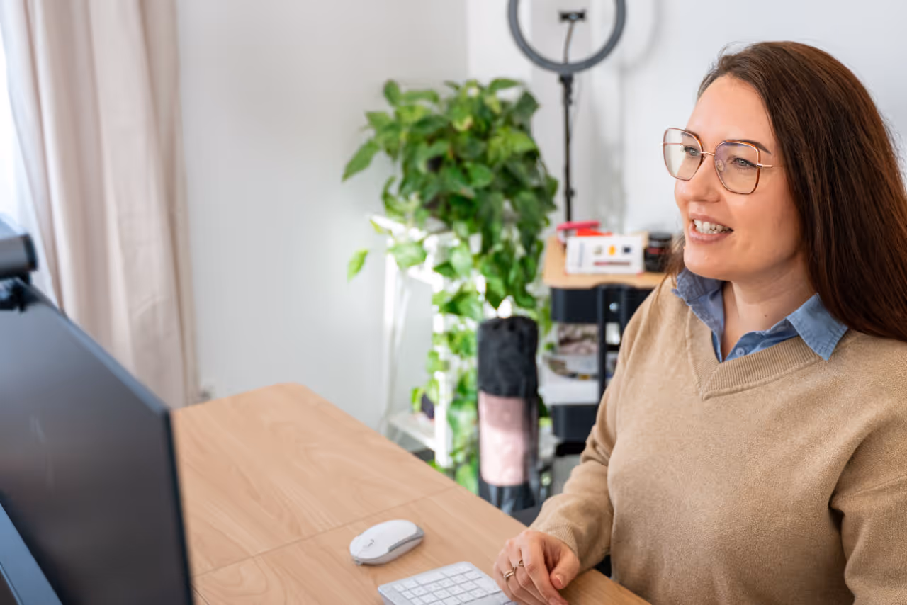 Frau mit Brille und braunem Pullover sitzt lächelnd vor einem Computerbildschirm in einem hellen Büro mit Pflanzen im Hintergrund.