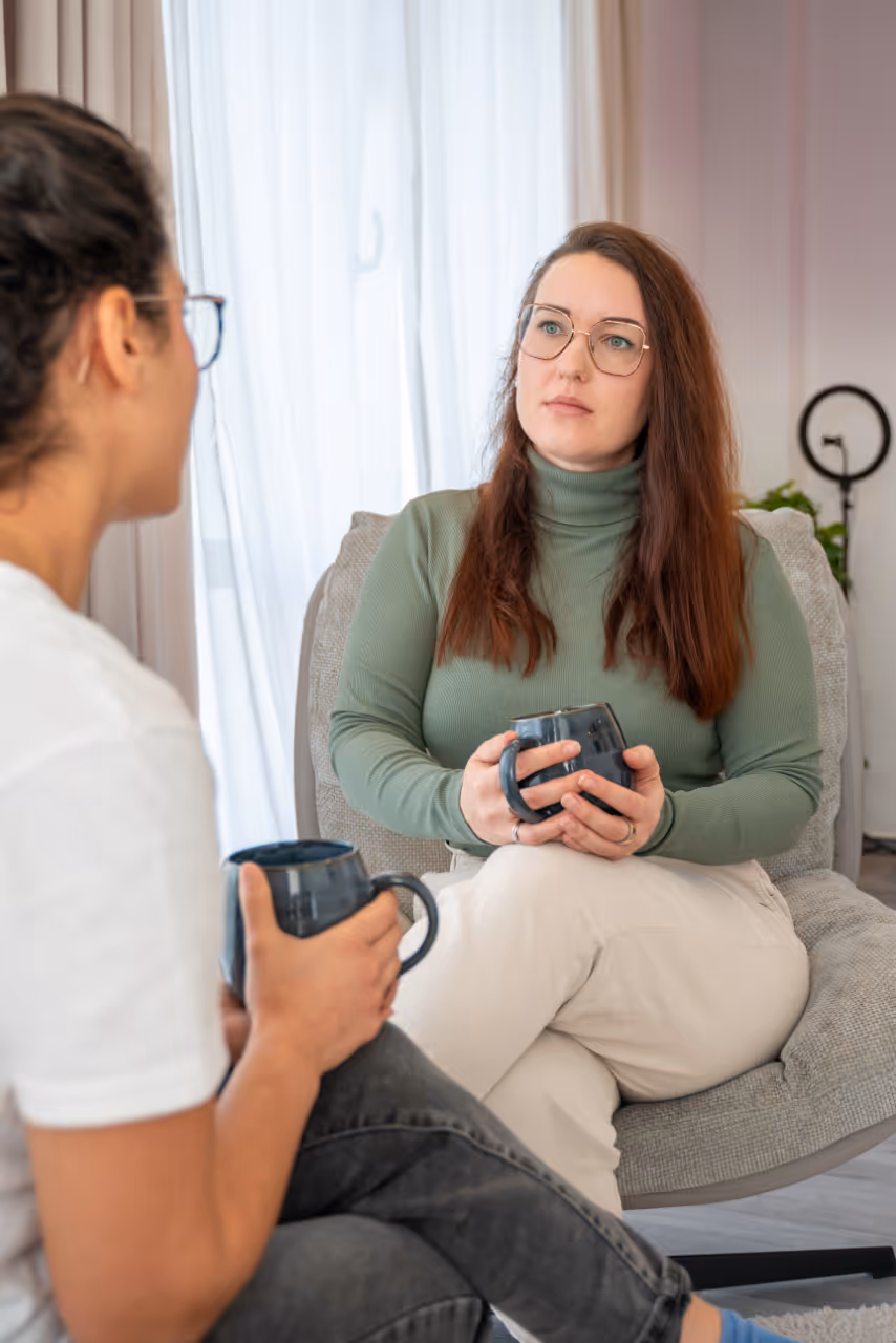 Zwei Frauen sitzen sich gegenüber und unterhalten sich bei einer Tasse Kaffee in einem gemütlichen Raum.