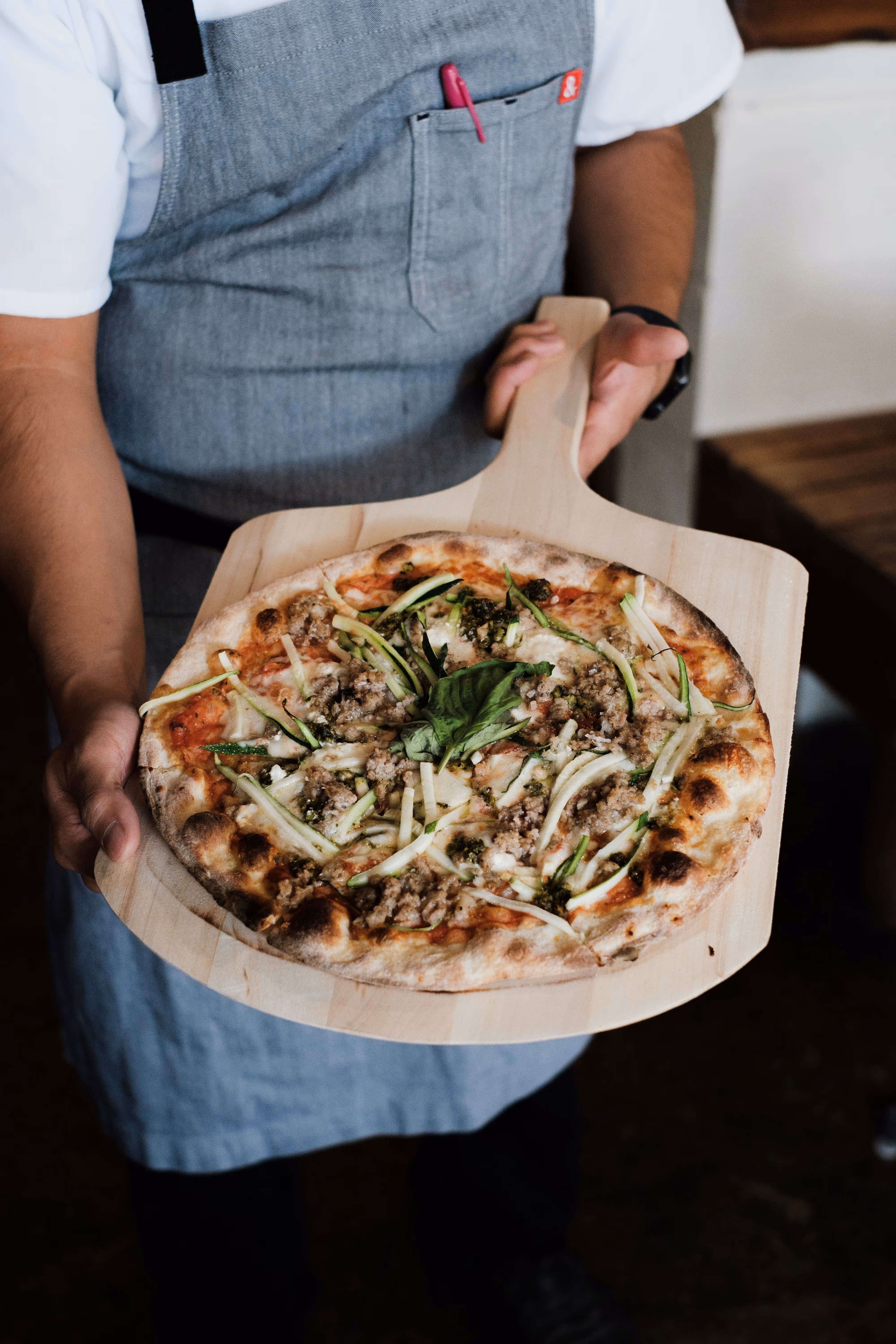 Person in a gray apron holding a wooden pizza peel with a freshly baked pizza topped with zucchini strips, ground meat, and basil leaves.