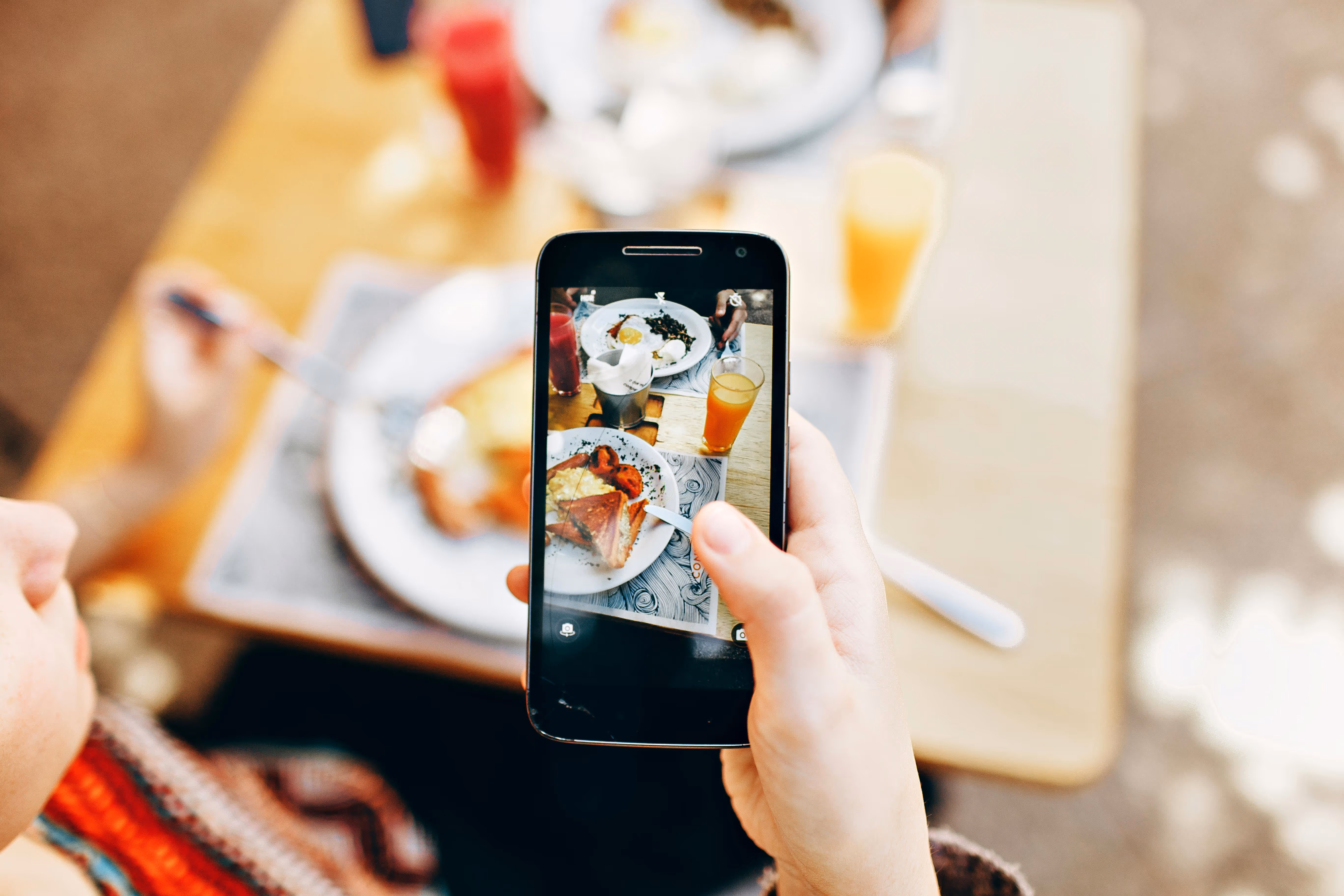 Close-up of a hand holding a smartphone capturing a photo of a breakfast plate with toast, eggs, and a glass of orange juice on a wooden table.