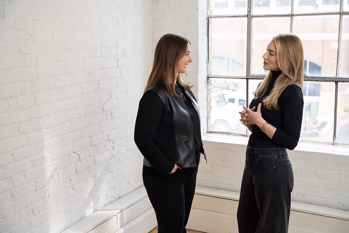 Two women standing and talking in a bright room with large windows and white brick walls.