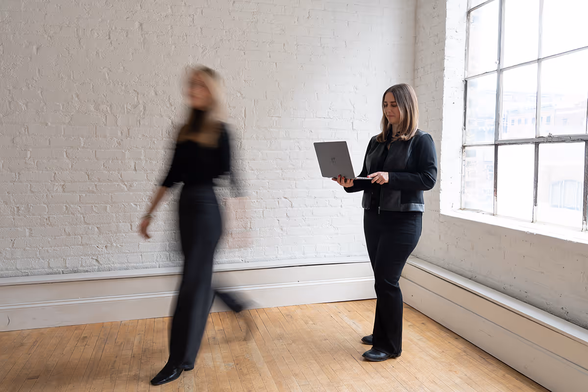 Woman in black jacket standing by a large window holding an open laptop while a blurred figure walks past her.