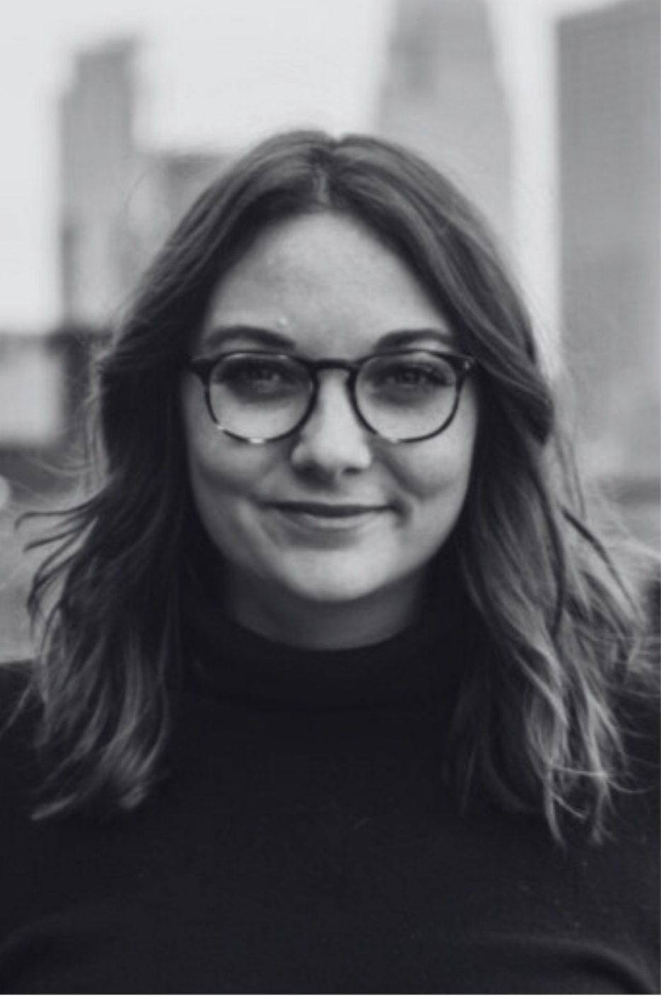 Black and white portrait of a young woman with shoulder-length hair, wearing glasses and a dark top, smiling slightly with a cityscape background.
