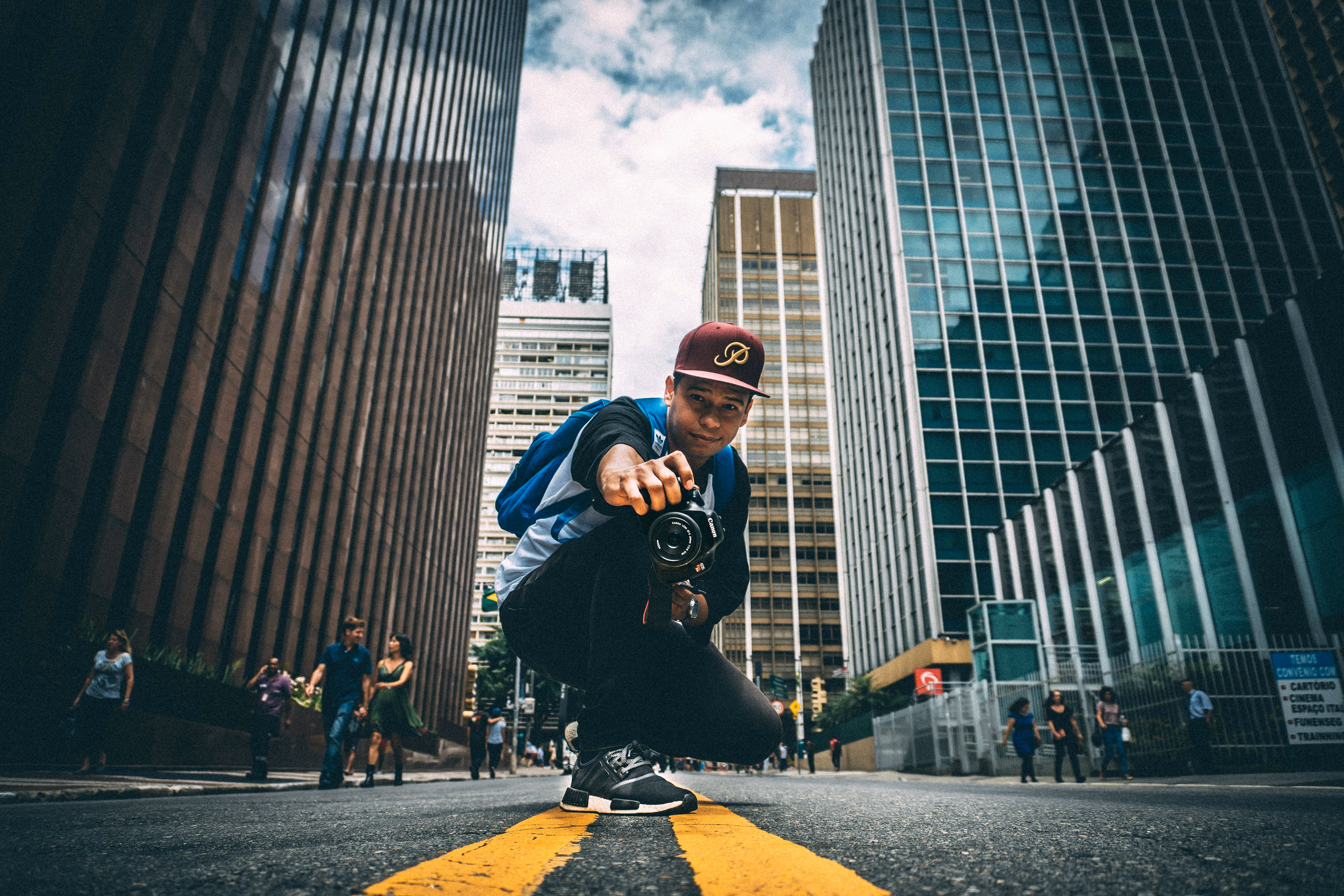 Young man crouching on a city street with a camera in hand, surrounded by tall buildings and pedestrians.