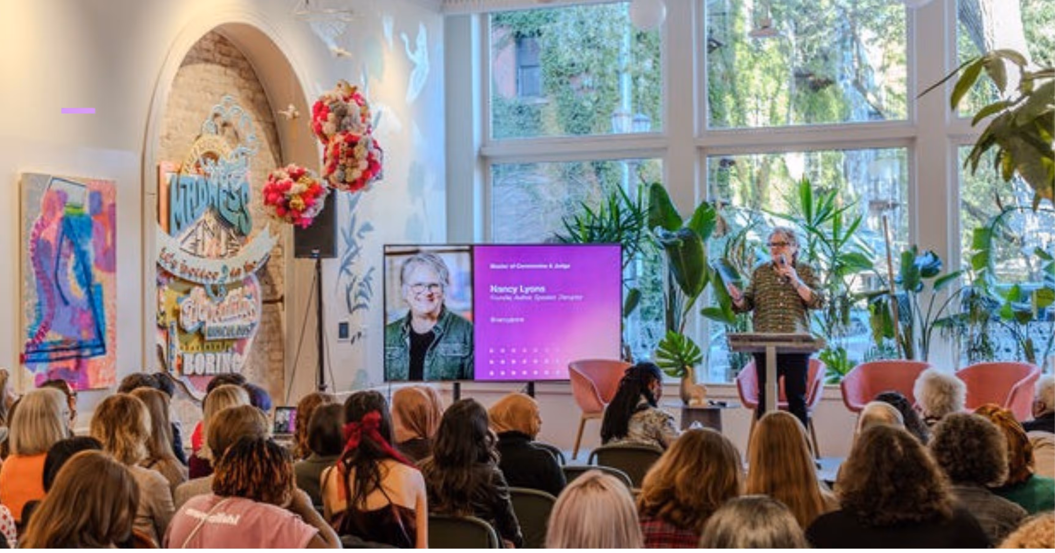 Speaker presenting in front of an audience in a bright room with large windows, plants, and a screen showing her photo and name, Nancy Lyons, Advisory Audio Design Program.
