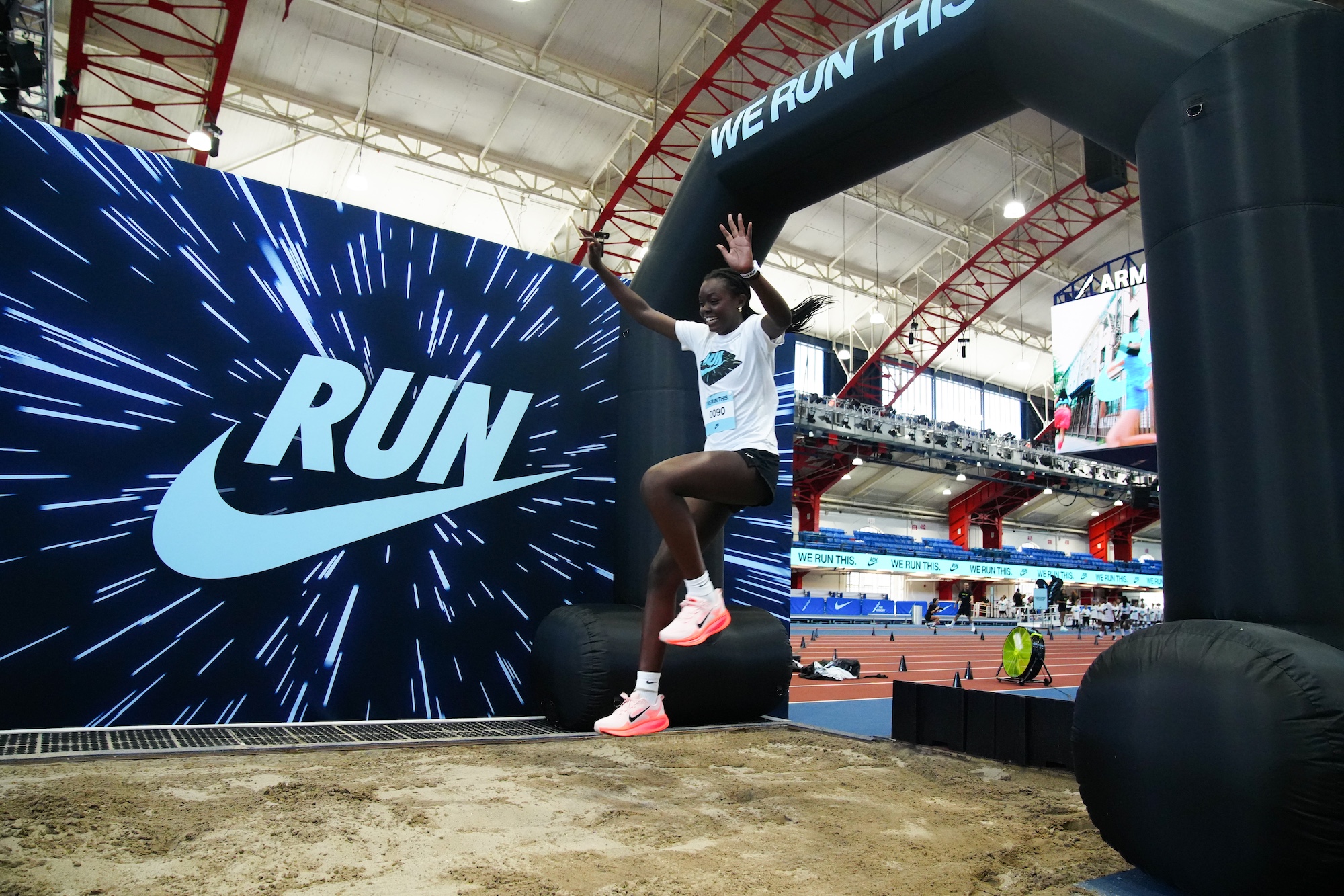 A girl smiles as she is midair, jumping into a track sand pit, a large Nike banner to her side.