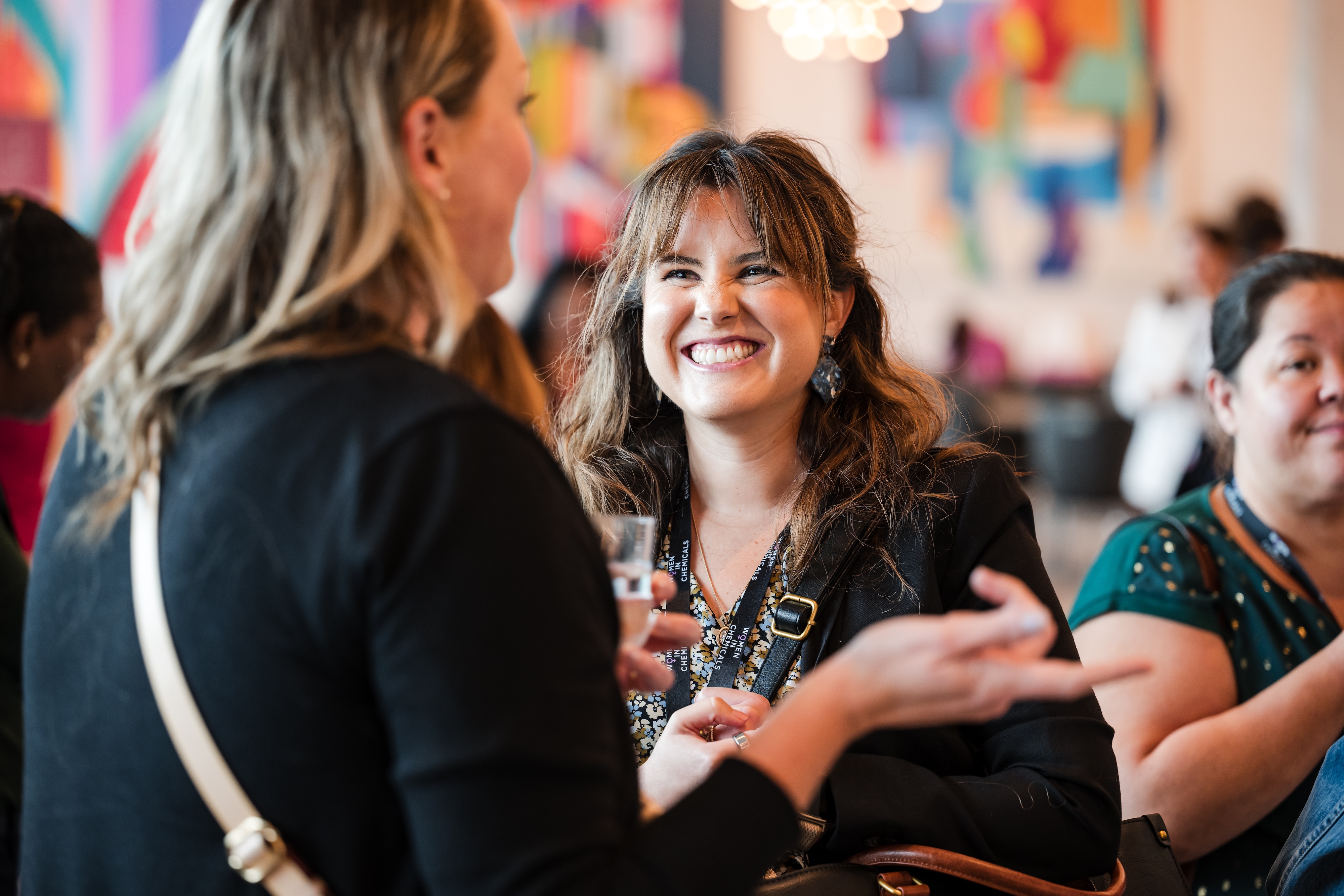 A woman wearing a blazer is smiling and laughing with the woman in front of her, in a crowded area.