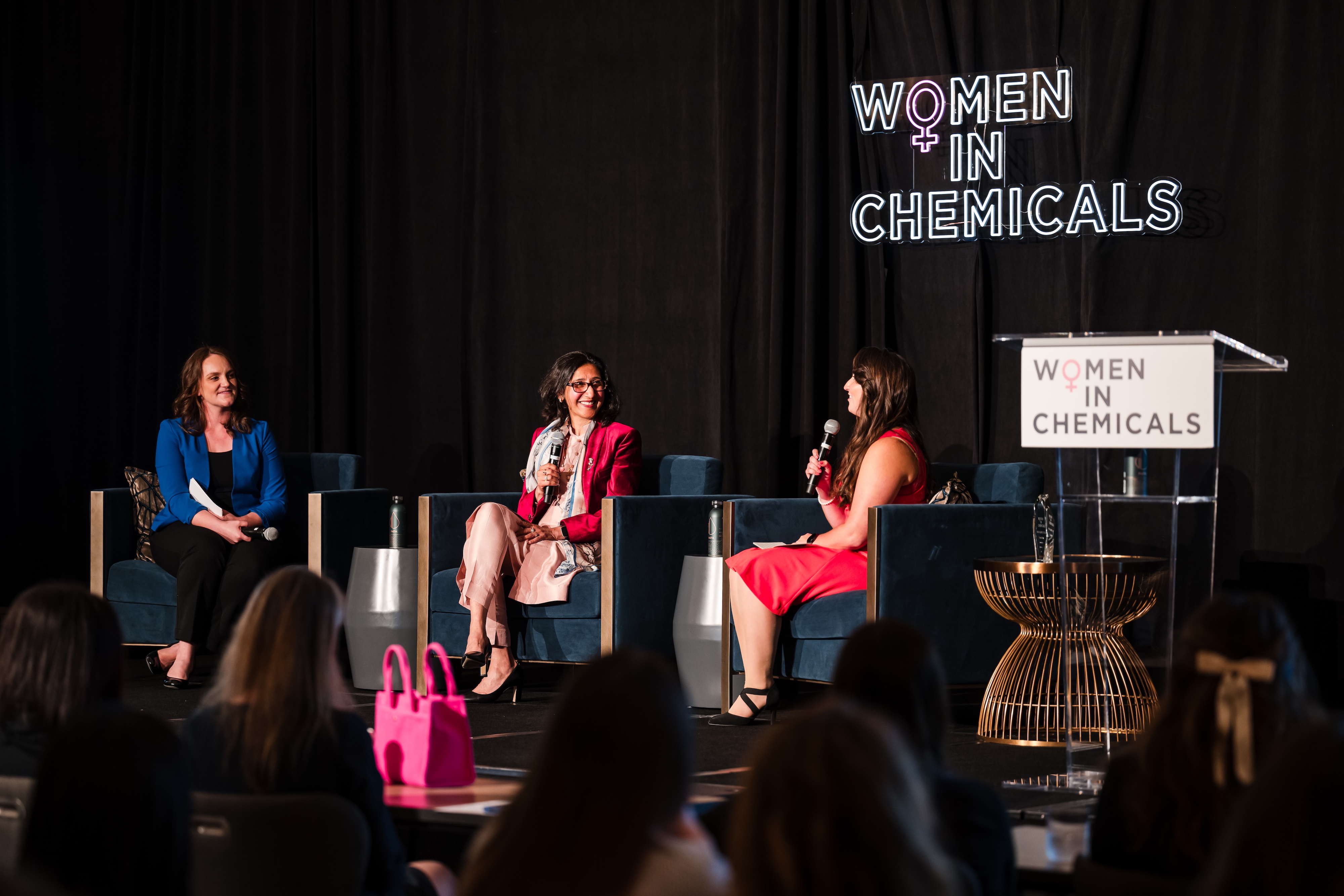 Three women sit on stage with microphones, a black curtain behind them with a neon sign saying "Women In Chemicals."