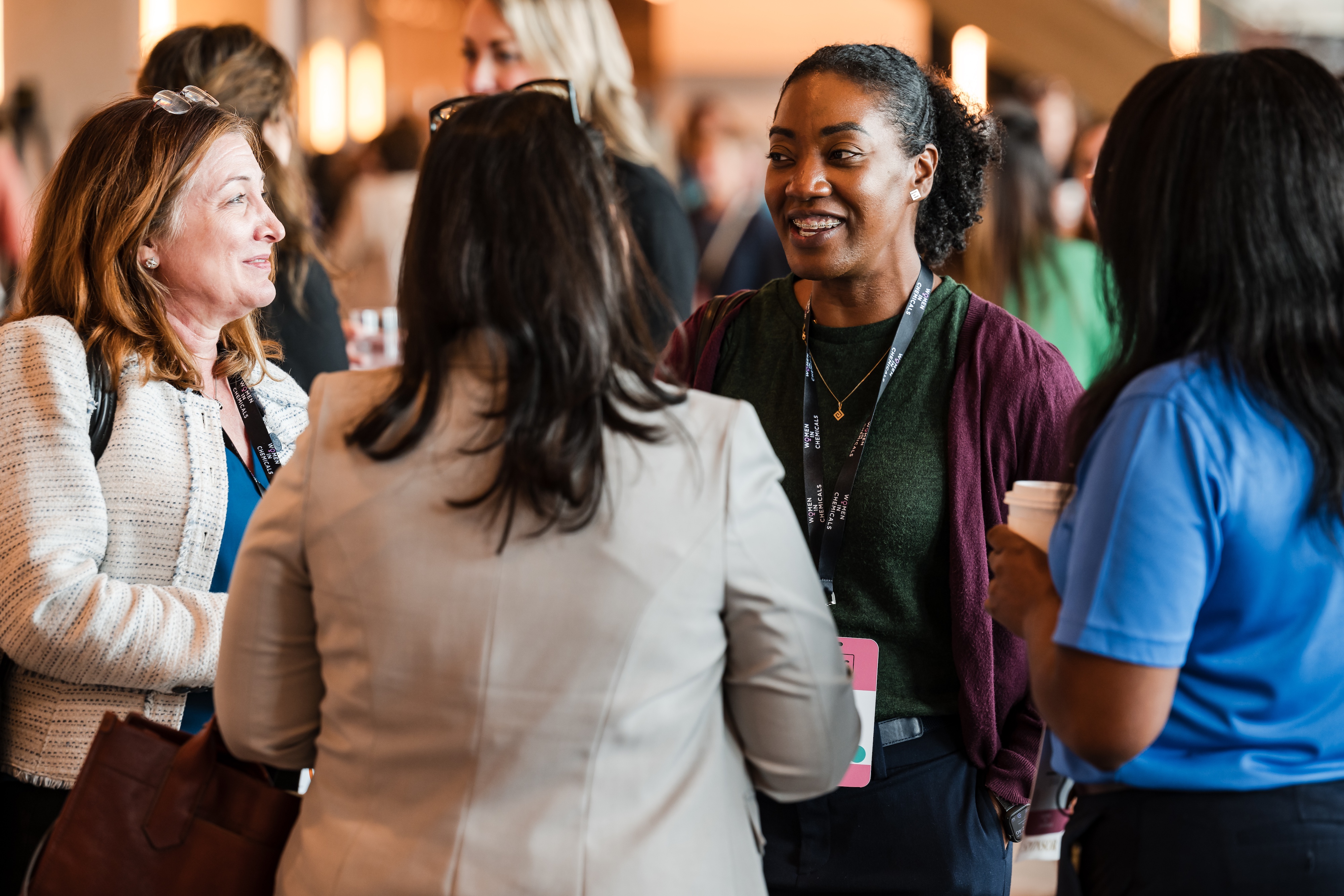 A woman wearing a purple sweater speaks with three others around her, and they wear lanyards saying "Women In Cheimcals."
