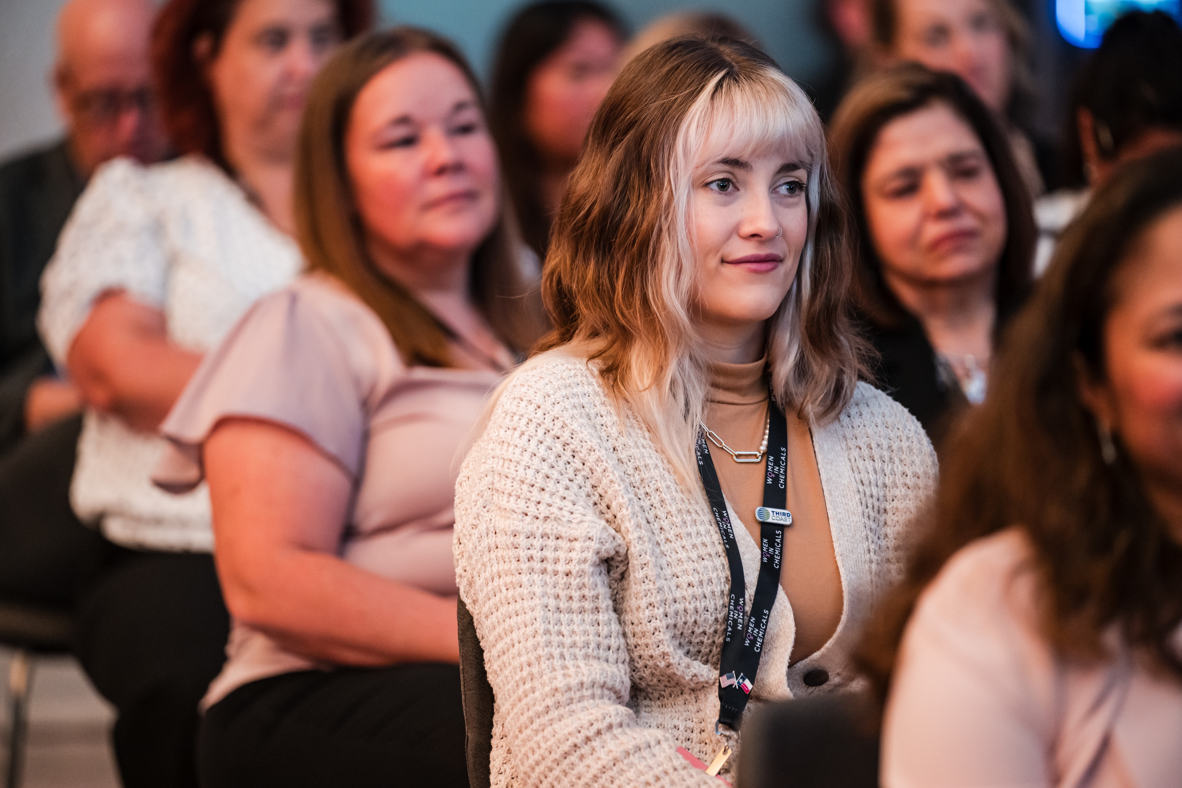 A woman wearing a white cardigan sits in the audience, listening to something in front of her.