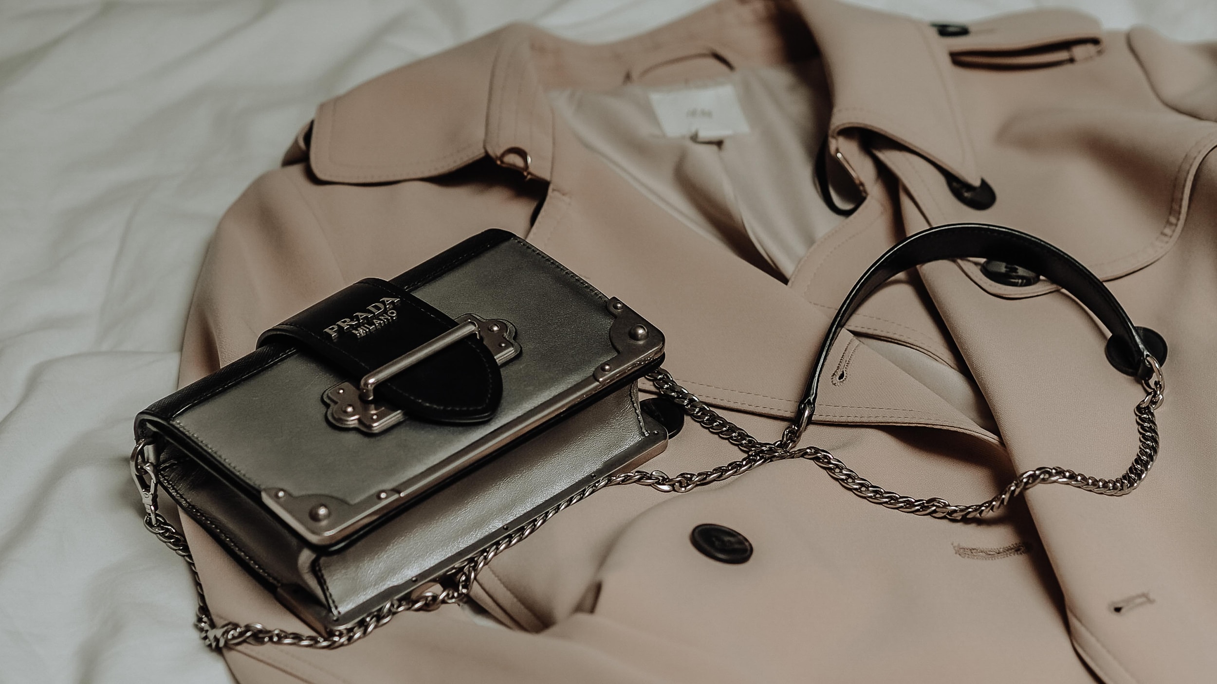A silver Prada bag is laid out over a trench coat on a white sheet.