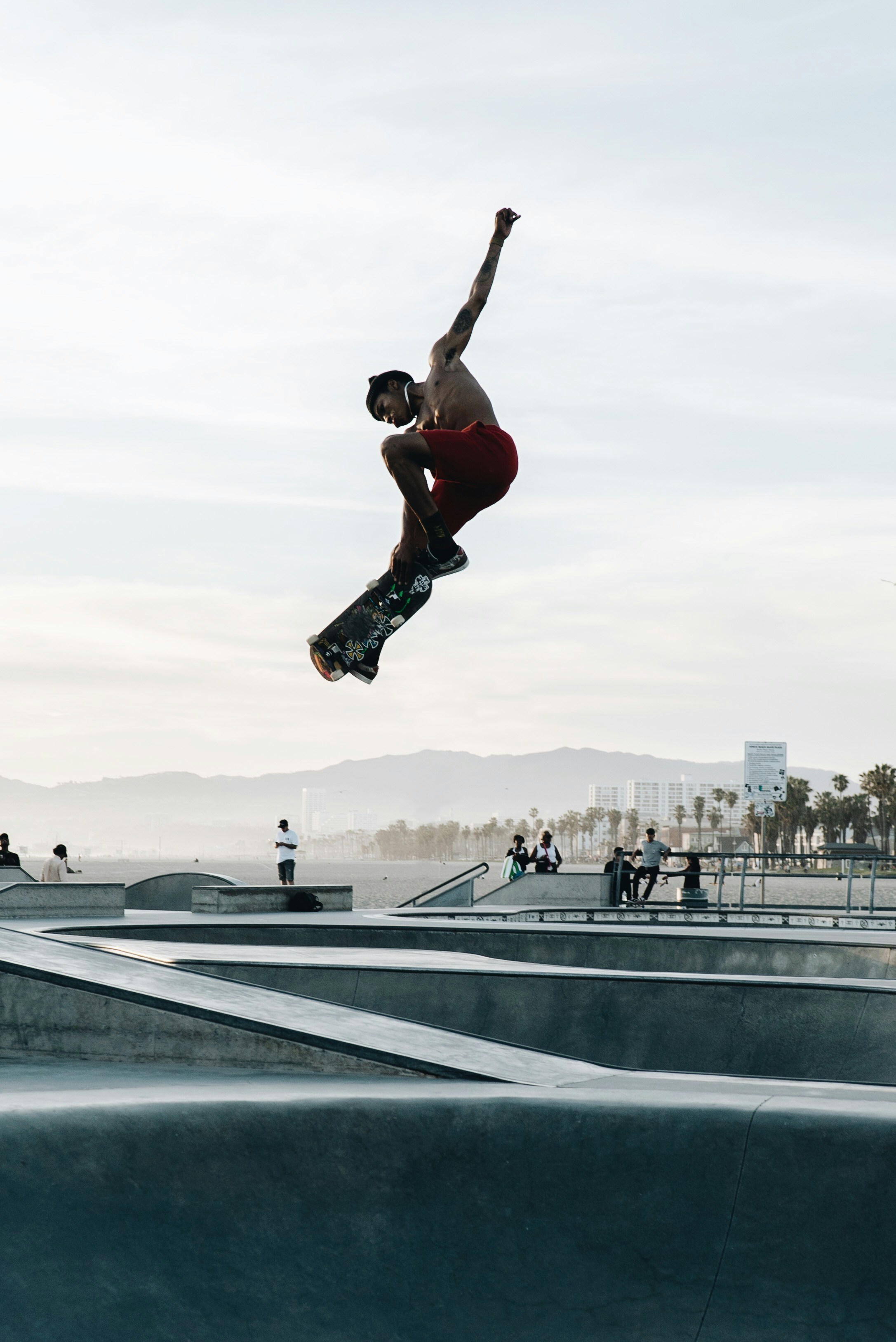 A skateboarder hovers midair over a ramp, somewhere along a beach with palm trees lining the sidewalk.