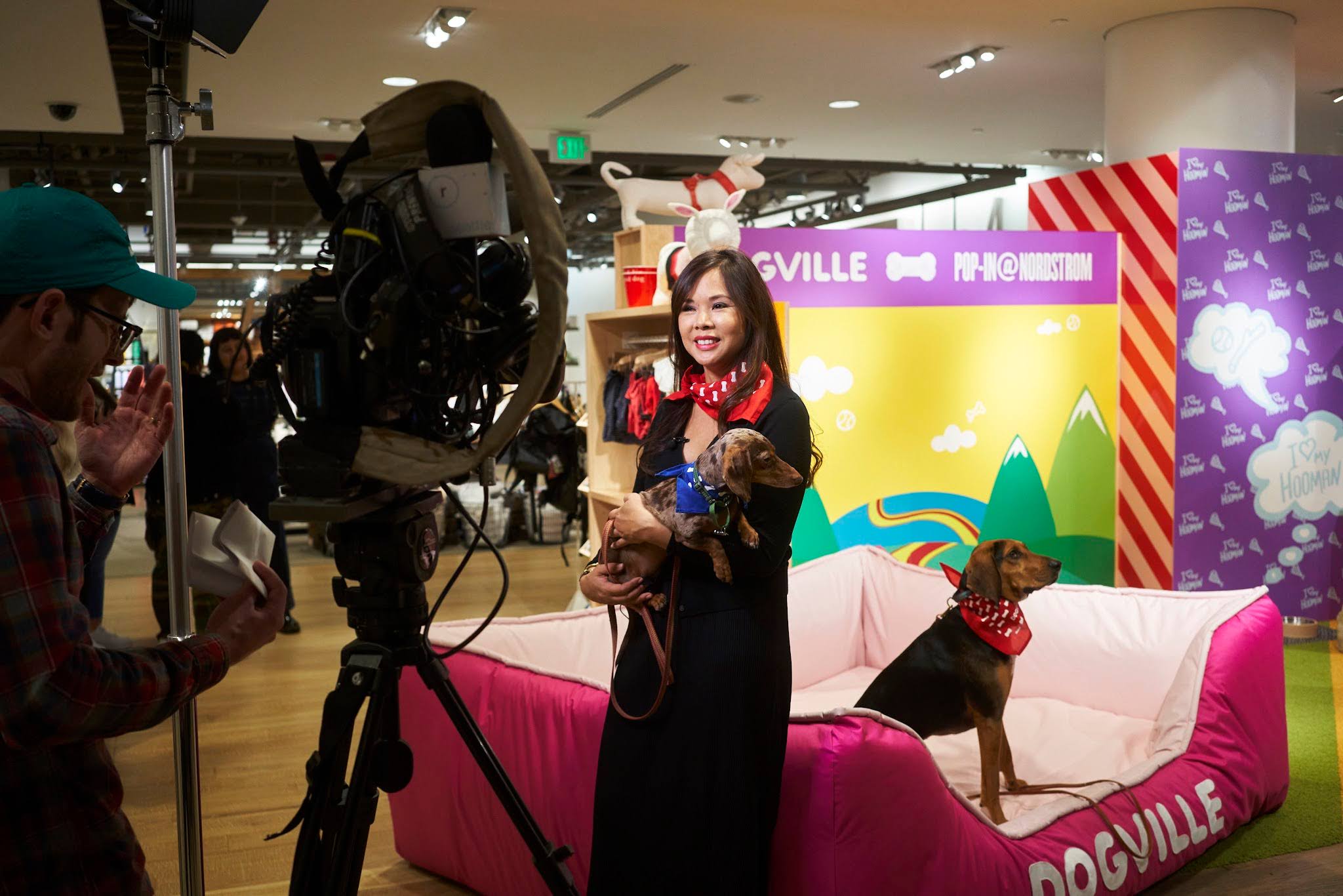 A woman holds a small dog in her arms while smiling at a camera, the Nordstrom Dogville pop-up sign in the background.