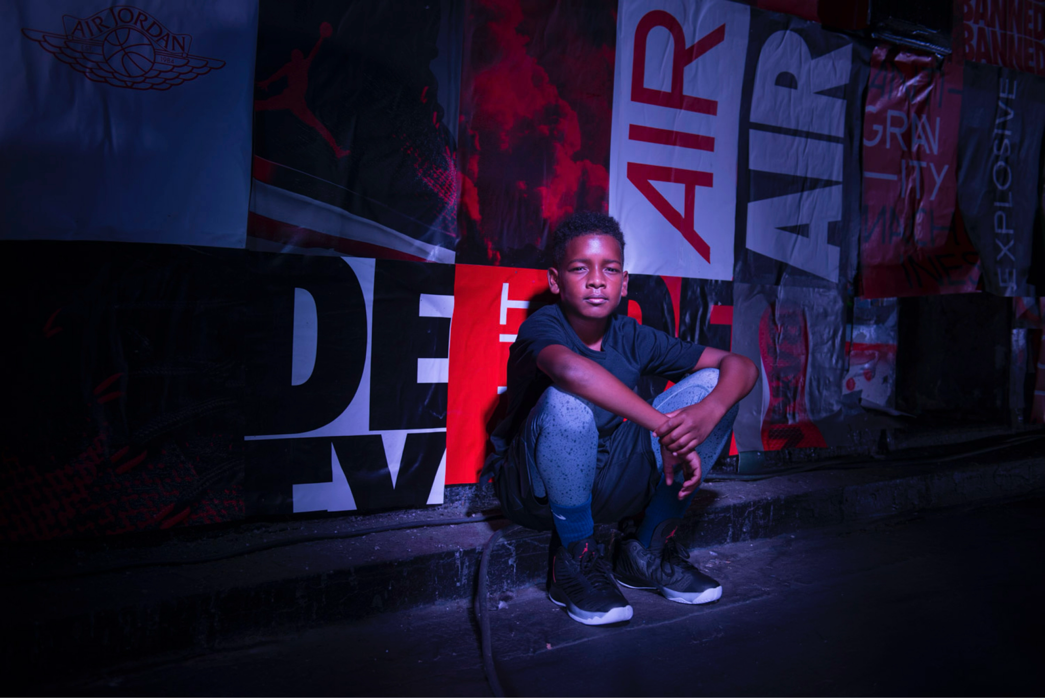 A young boy sits in front of a wall plastered in different Jordan posters.