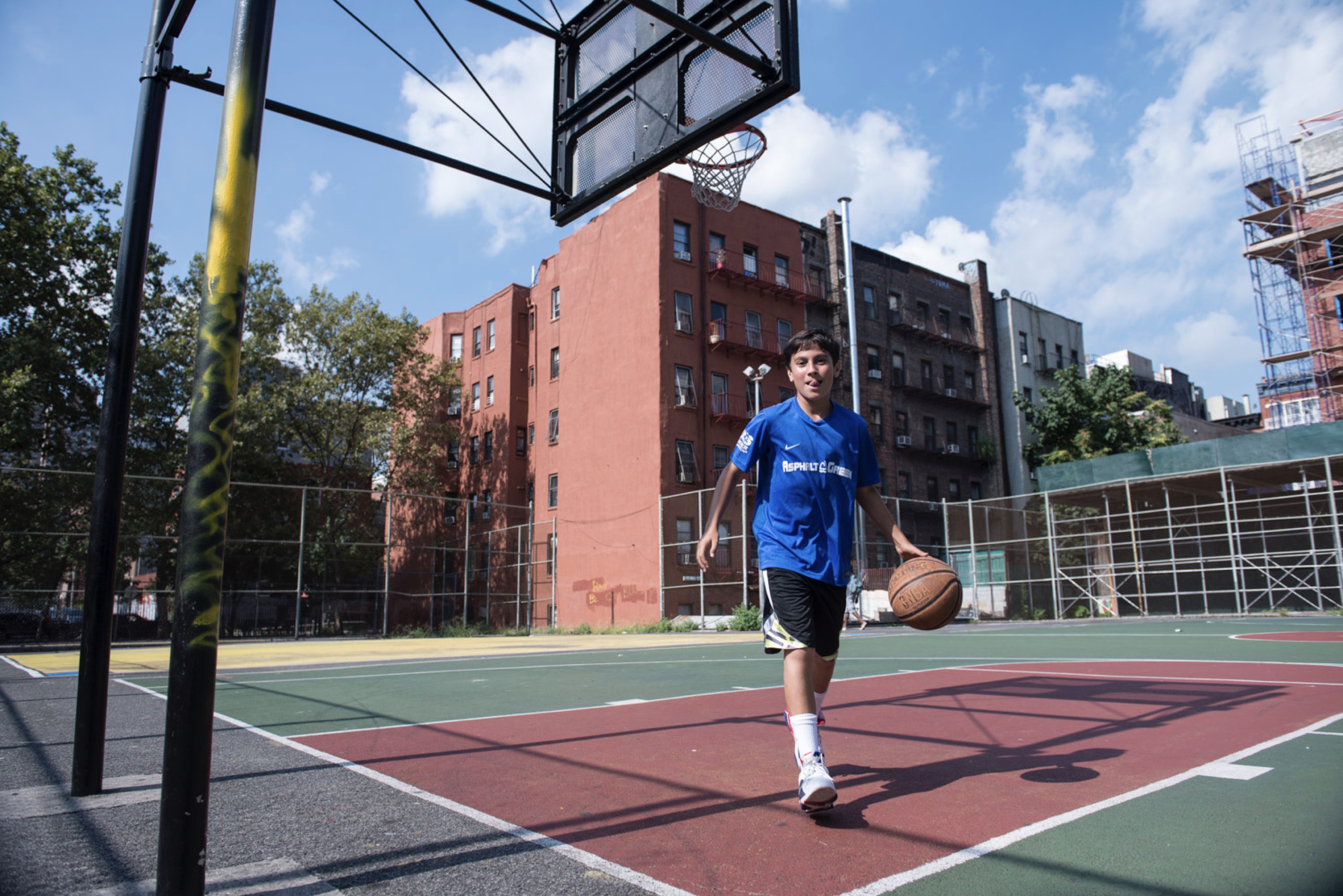 A boy walks towards the camera, dribbling a basketball on an outside court.