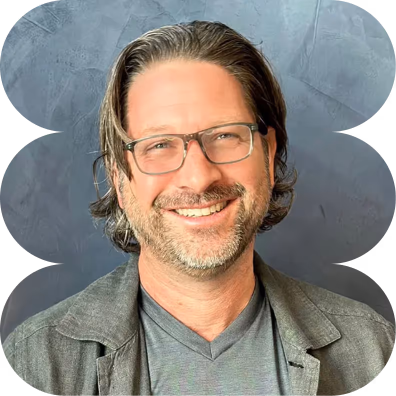 Smiling man with shoulder-length brown hair, glasses, and a beard wearing a gray jacket and t-shirt against a textured gray background.