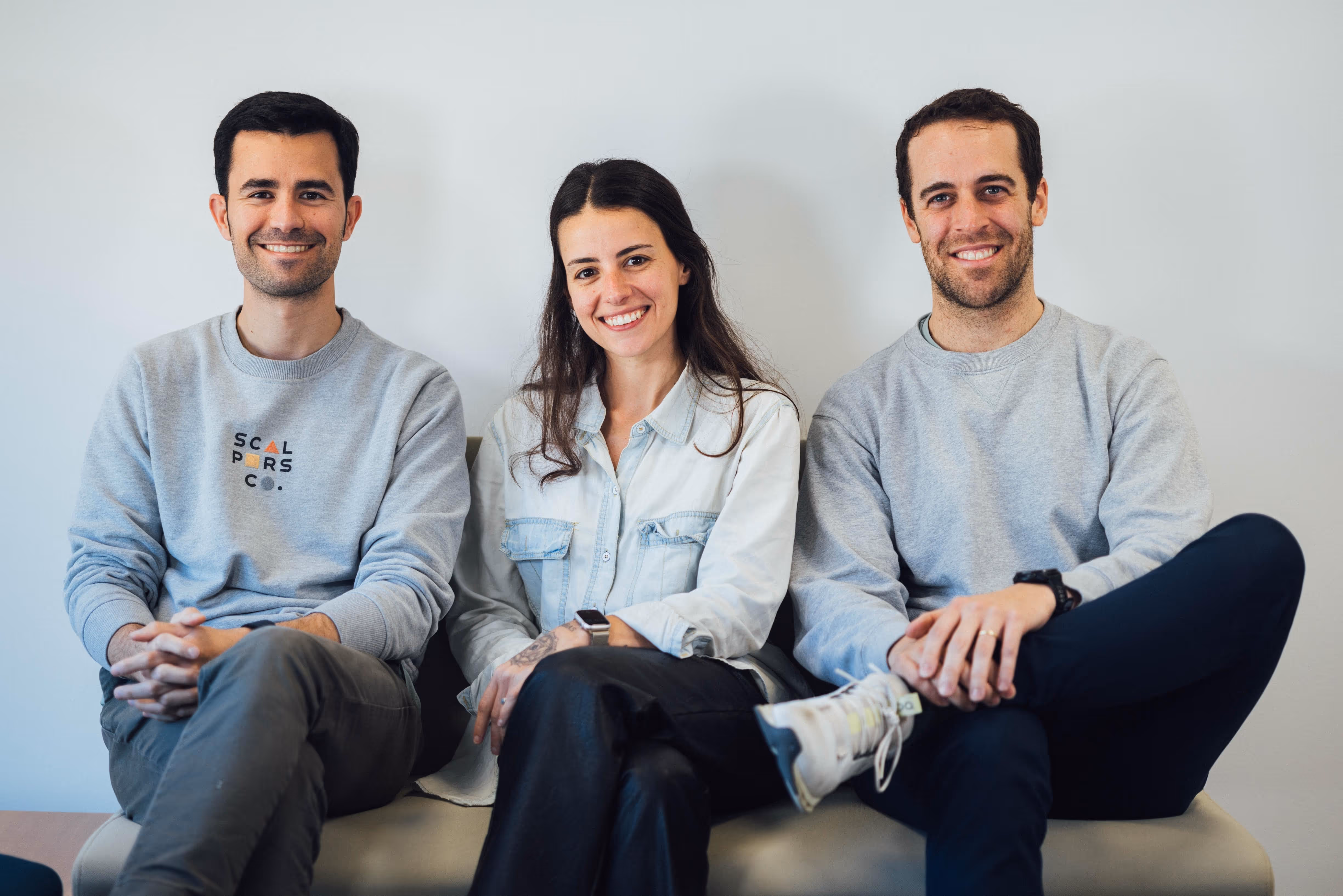 Three young adults sitting on a bench against a plain white wall, smiling at the camera.
