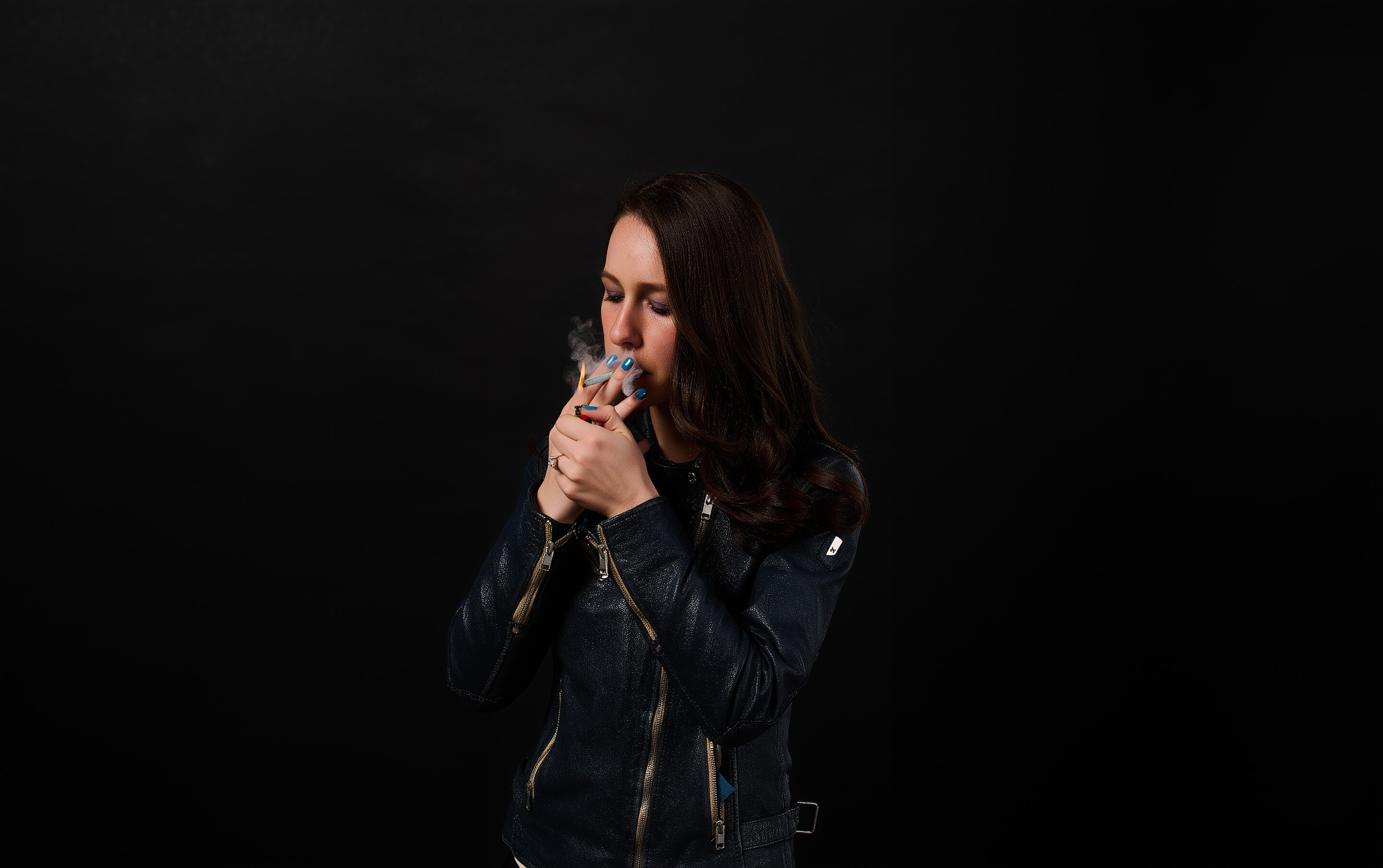 Woman with long dark hair wearing a black leather jacket lighting a cigarette against a black background.