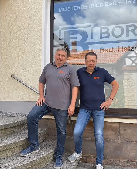 Zwei Männer in Jeans und Polo-Shirts lehnen vor einem Fenster mit der Aufschrift ‚MEISTERBETRIEB BAD + HEIZ‘.