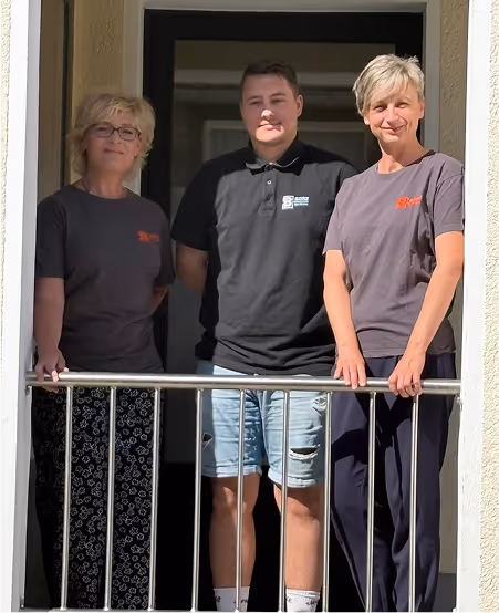 Drei Personen, zwei Frauen und ein Mann, stehen auf einem Balkon vor einer Tür und blicken in die Kamera.