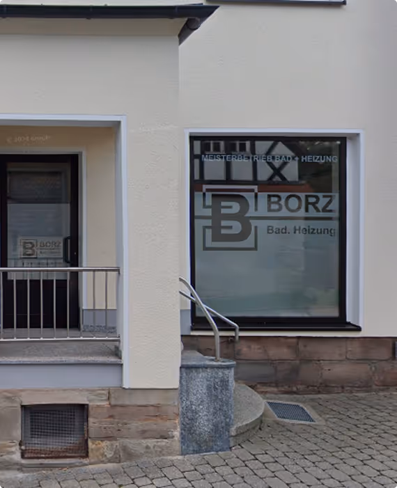 Fassadenansicht eines Gebäudes mit Eingangstür und großem Fenster mit Beschriftung 'MEISTERBETRIEB BAD + HEIZUNG BORZ Bad. Heizung.'