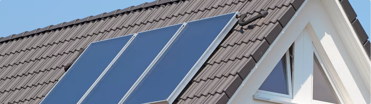 Drei Solarpanels auf dem Dach eines Hauses mit braunen Ziegeln und weißem Giebelfenster.