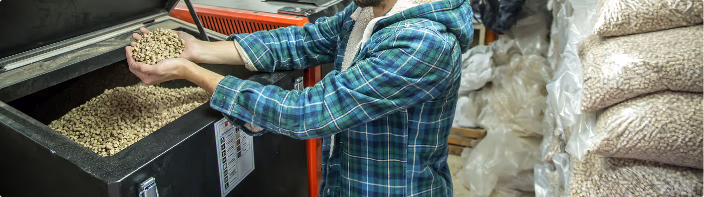 Person hält Holzpellets mit beiden Händen über einem offenen Behälter, daneben Stapel von gefüllten Plastiksäcken mit Holzpellets.