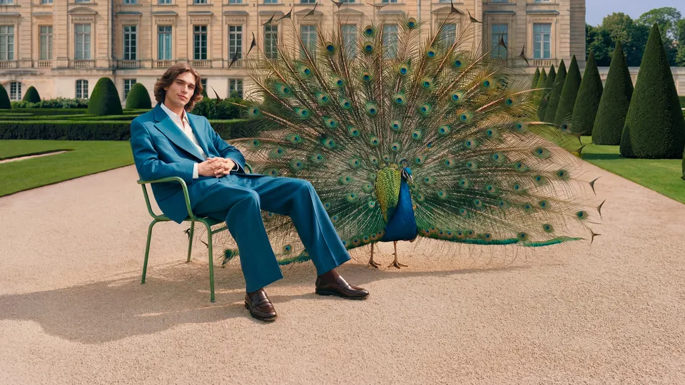 Man in teal suit seated before a chateau with a peacock displaying its feathers, Parfums de Marly — luxury video production Detroit, photography & DA by Alex