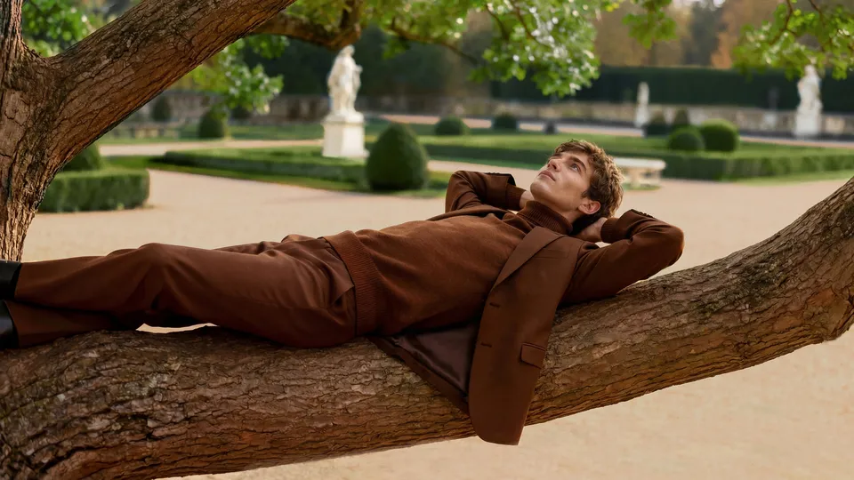 Man in brown tailored knitwear reclining on a tree branch in a formal garden, Parfums de Marly — luxury video production Detroit, photography & DA by Alex