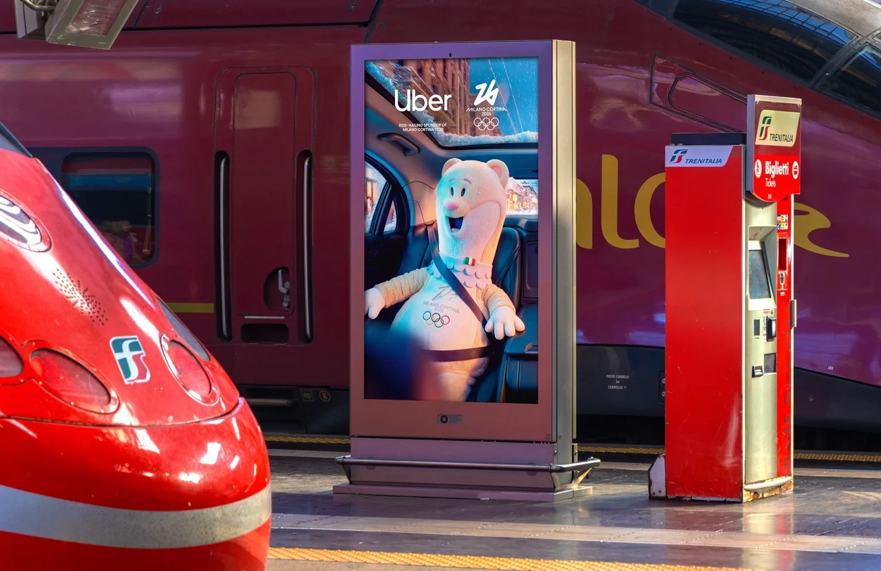 Digital kiosk screen beside a Trenitalia train showing the Uber Milano Cortina 2026 mascot secured in a car seat — 3d product visualization Detroit, photography & art direction by Detroit AI Production House
