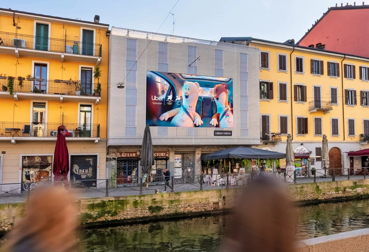 Digital LED billboard on a canalside Milan building showing the Uber Milano Cortina 2026 mascots at dusk — virtual production studio Detroit, photography & art direction by Detroit AI Production House