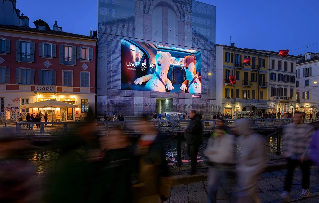 Illuminated Uber Milano Cortina 2026 screen glowing above a night piazza as blurred pedestrians pass below — ai video production Detroit, photography & art direction by Detroit AI Production House
