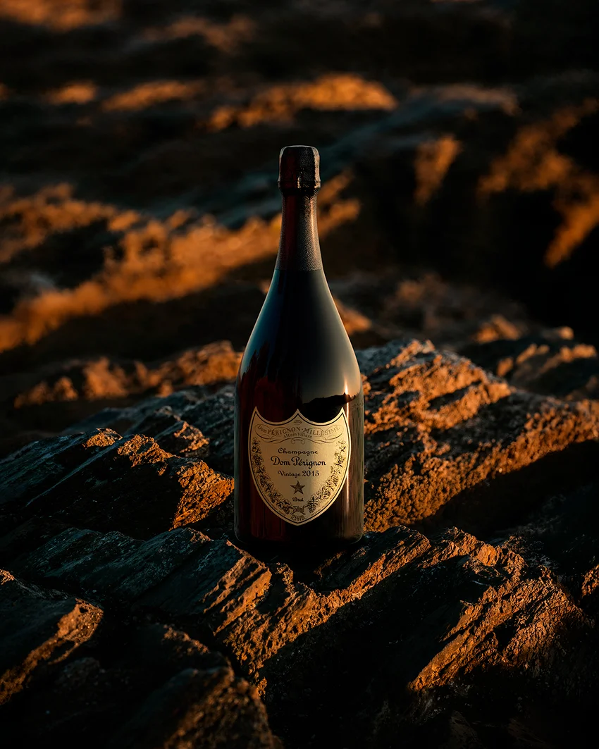 Dom Pérignon bottle standing on sunlit rocks at golden hour with the sea stretching behind — virtual production studio Detroit, photography & art direction by Pierrick Jégou, produced by Detroit AI Production House