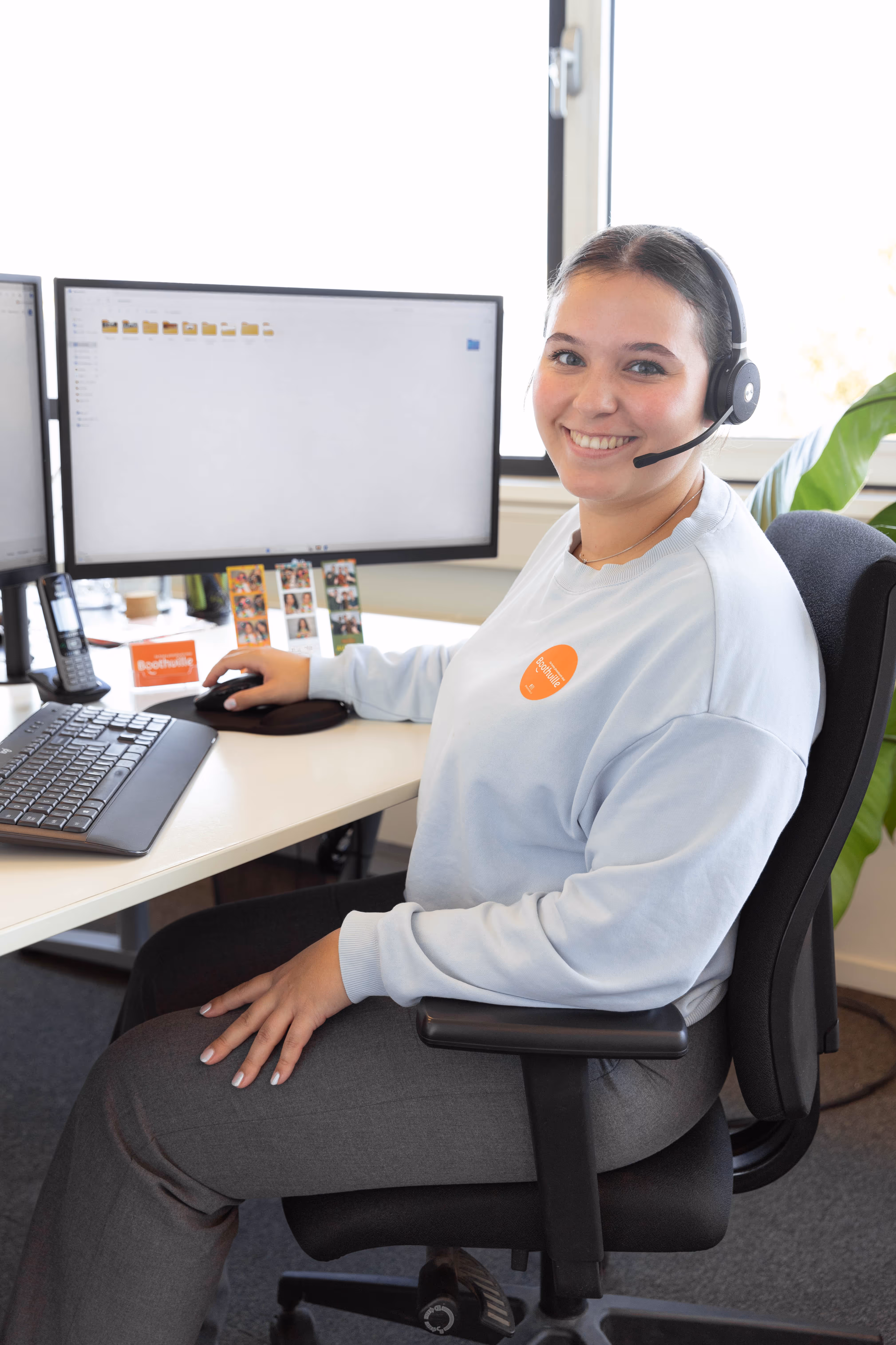 Vrouw met headset werkt zittend aan bureau met twee computermonitoren en toetsenbord.