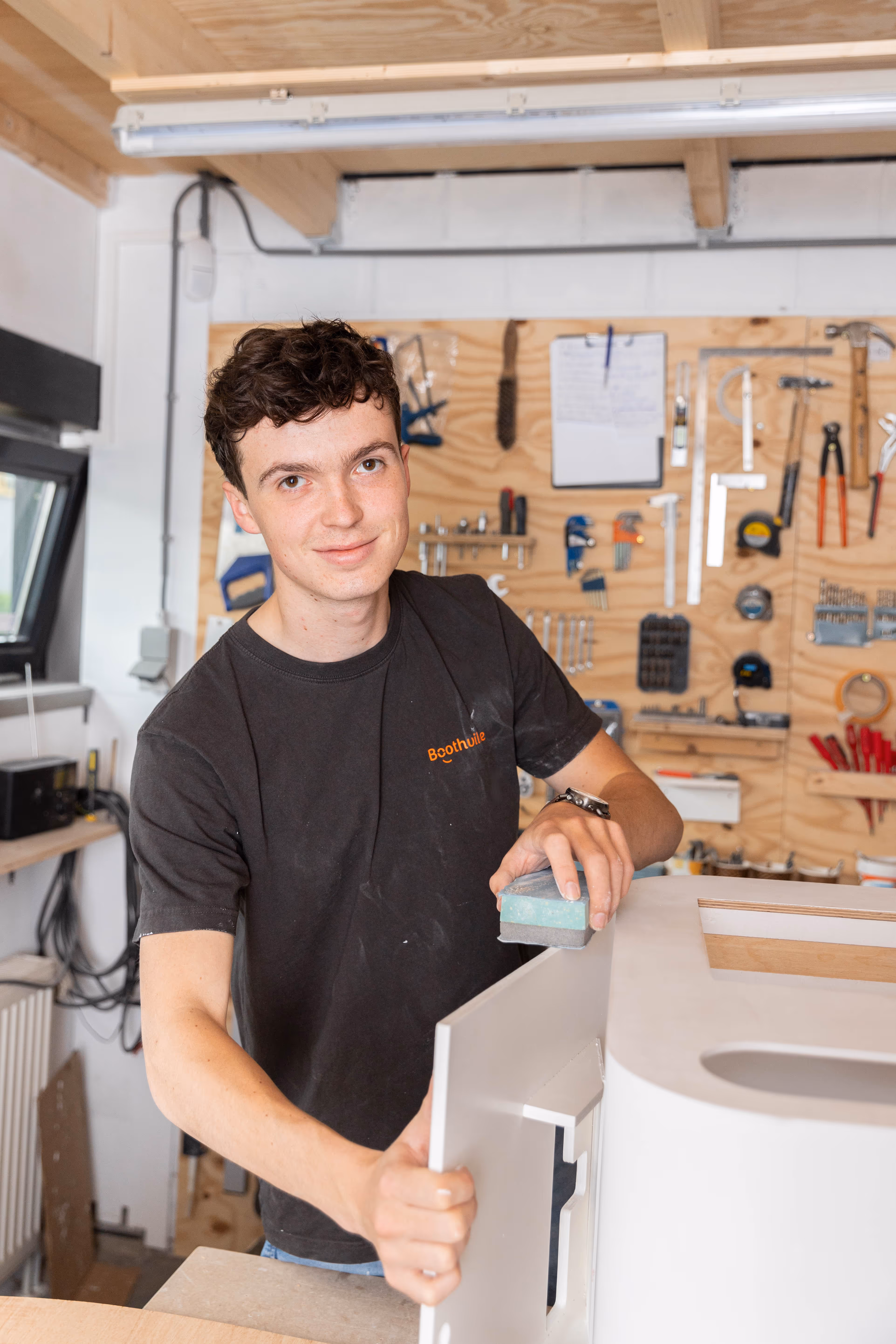Technisch uitvoerder van Boothville schuurt een wit photobooth deel in een werkplaats met diverse gereedschappen aan de muur.