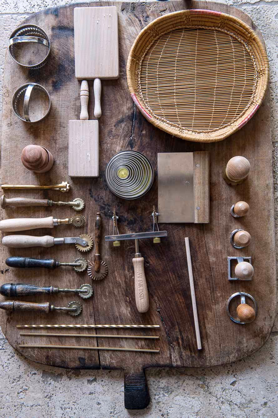 Varietà di utensili da cucina tradizionali e strumenti artigianali disposti ordinatamente su un tagliere di legno rustico.