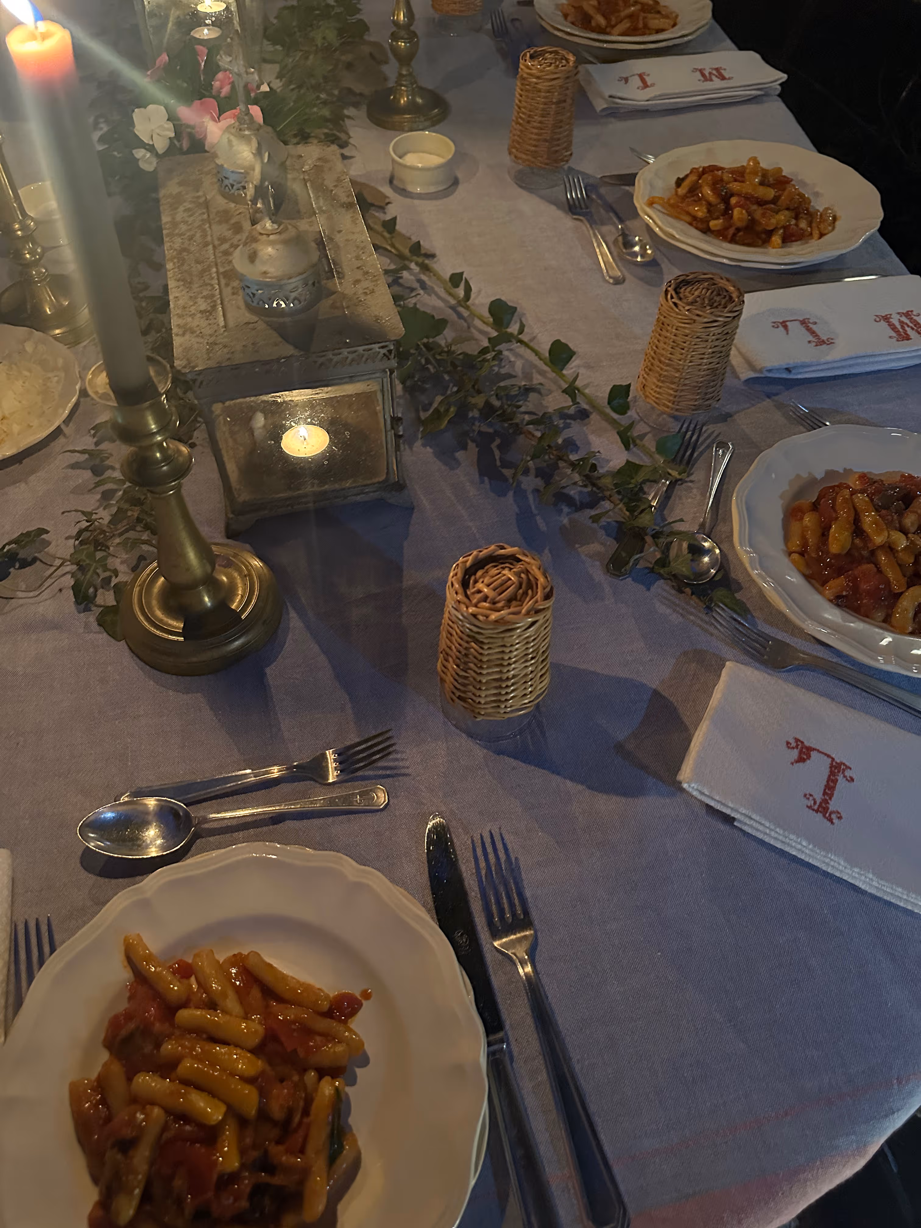 Tavola apparecchiata con piatti di pasta al sugo, candela accesa e centrotavola con foglie verdi su tovaglia grigia.