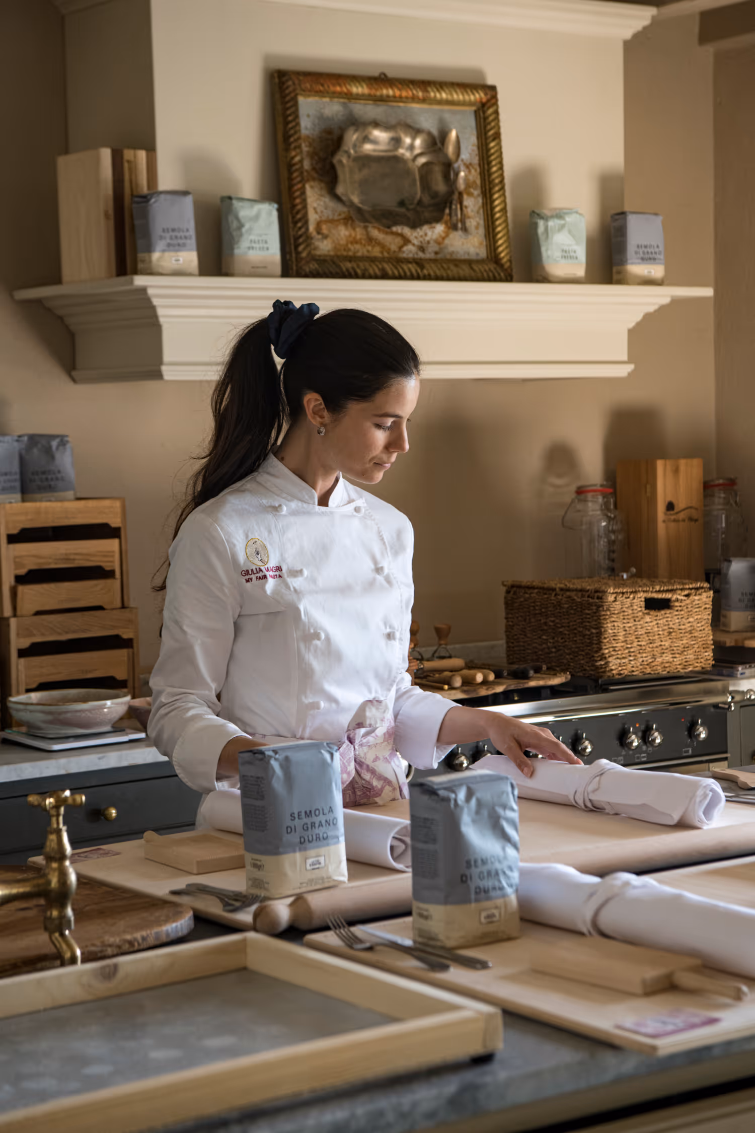 Donna in giacca da chef bianca prepara una lezione di pasta in cucina rustica con sacchetti di semola di grano duro sul tavolo.