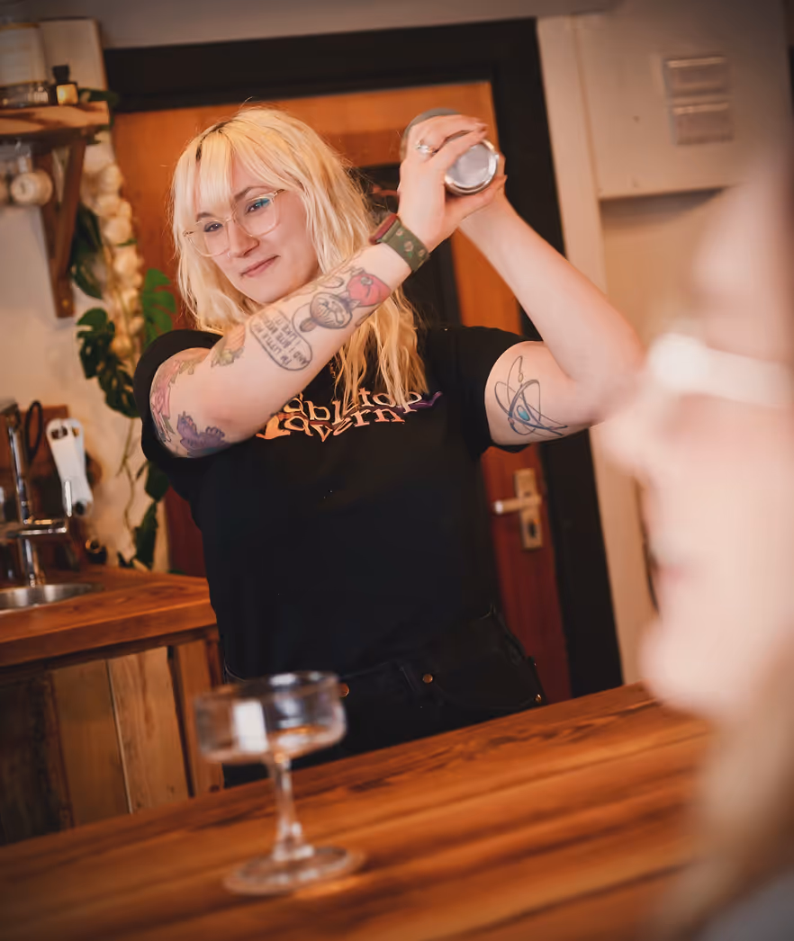 Person with blonde hair and tattoos shaking a cocktail shaker behind a wooden bar counter.