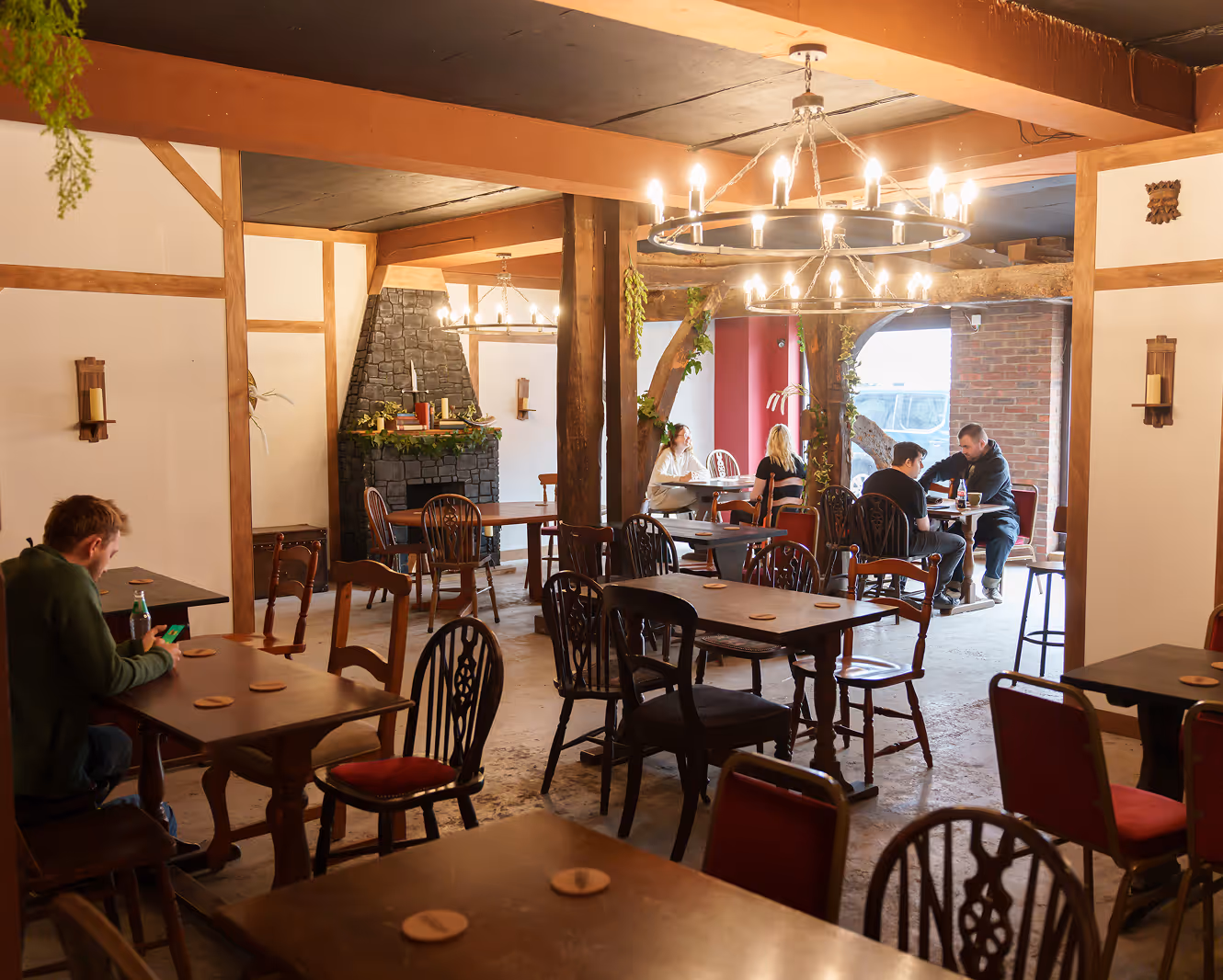 Cozy rustic cafe interior with wooden tables and chairs, chandeliers, a stone fireplace, and patrons seated at various tables.